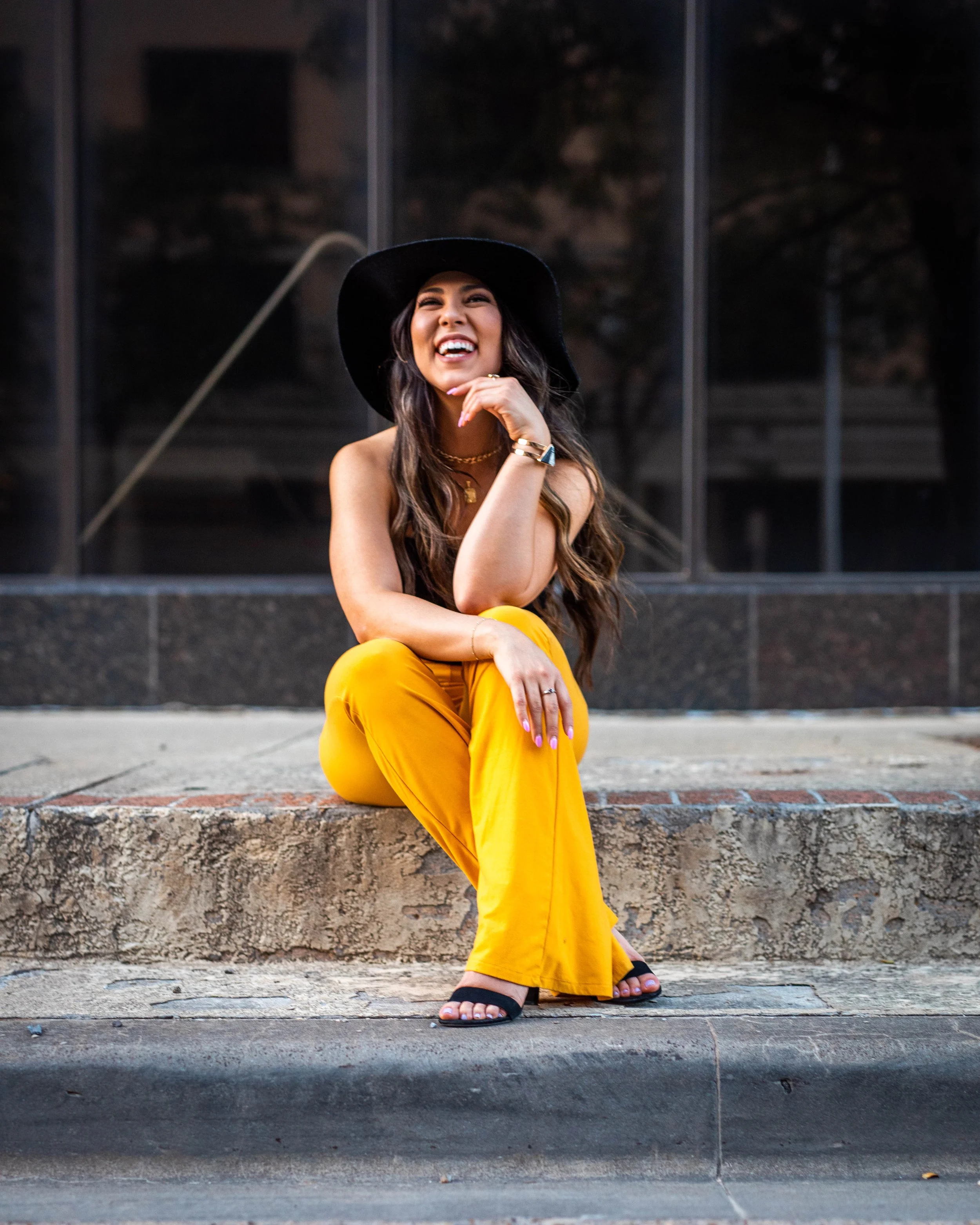 A woman sitting on a sidewalk in front of a glass building, wearing a black hat, yellow pants, black sandals, and smiling while looking away.