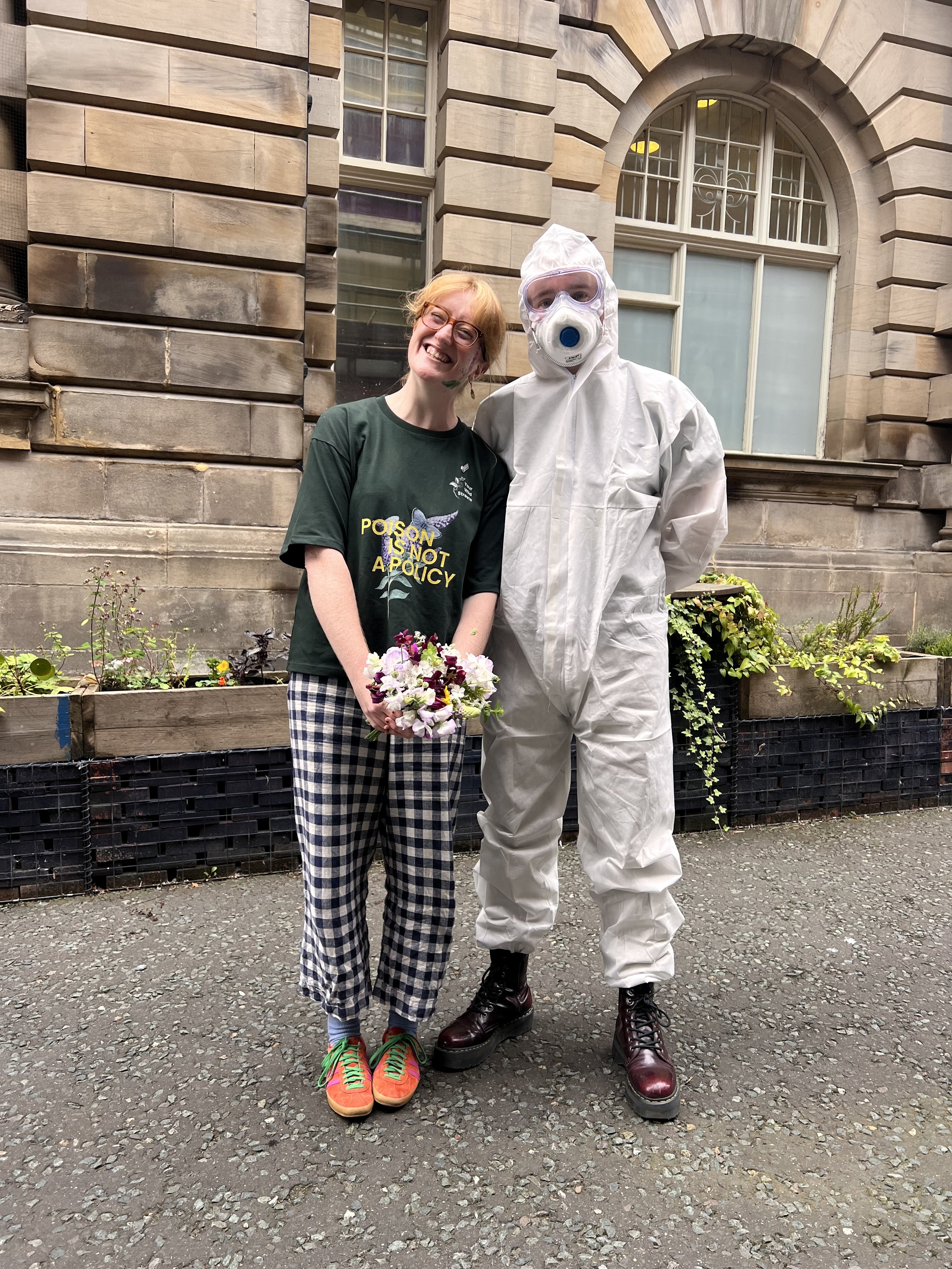Roisin stands with a man in full PPE suit, holding a bunch of sweet peas.