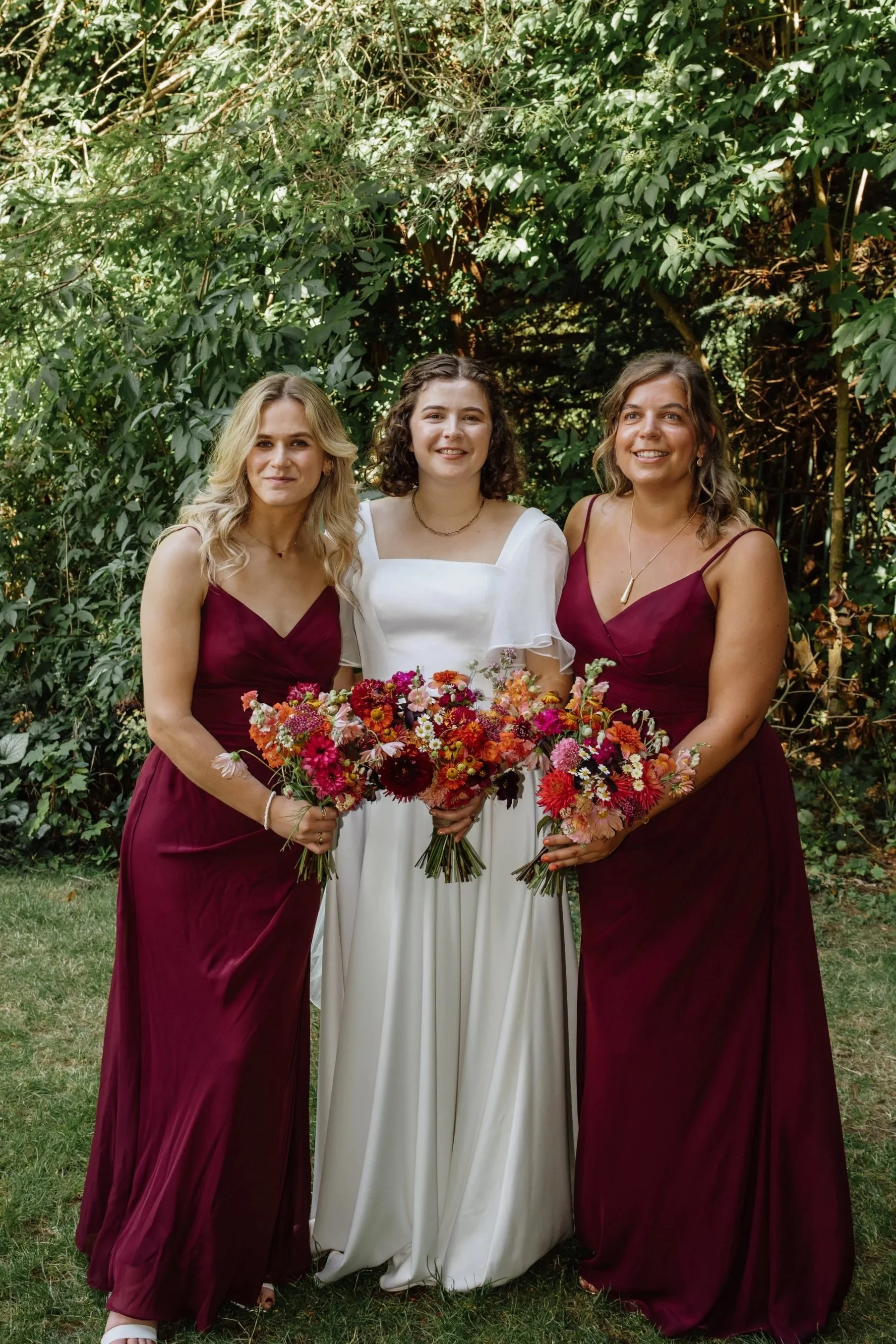Three white women, the bride in white in the centre, with two women in red bridesmaids dresses to her left and right hold their bouquets in the centre of the image. The bouquets are mostly red, orange and white flowers.
