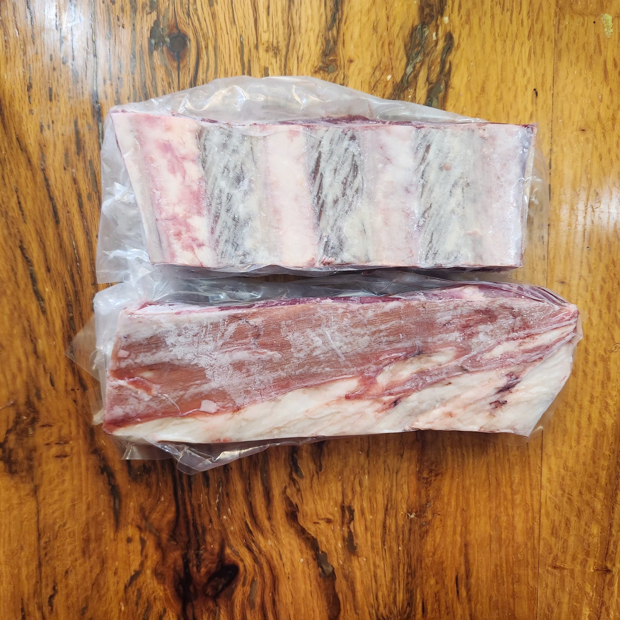 Two packages of beef short ribs in packages on brown table flipped upside down to see underside.