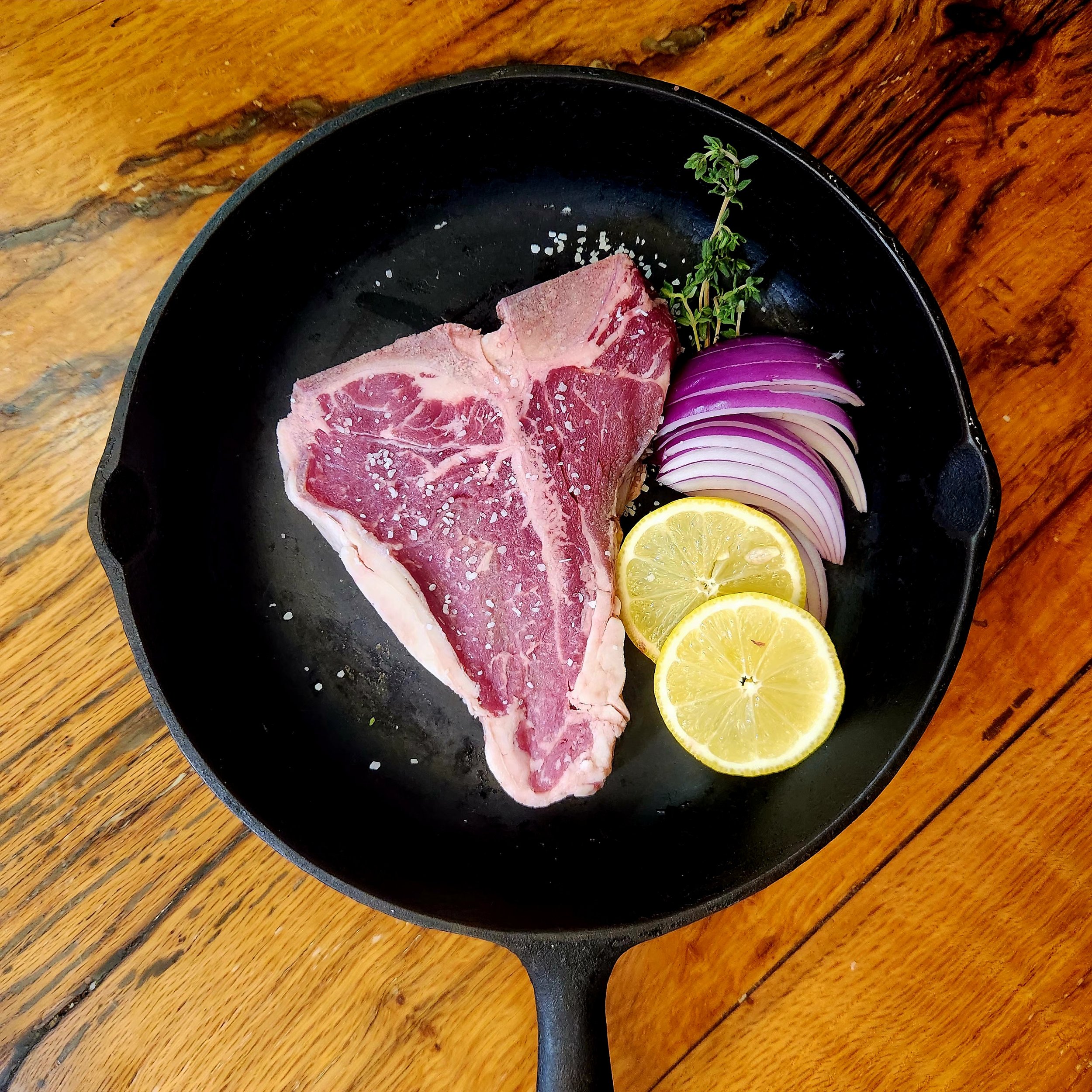Beef Porterhouse steak. In black cast iron pan with herbs and seasonings. Placed on brown table.