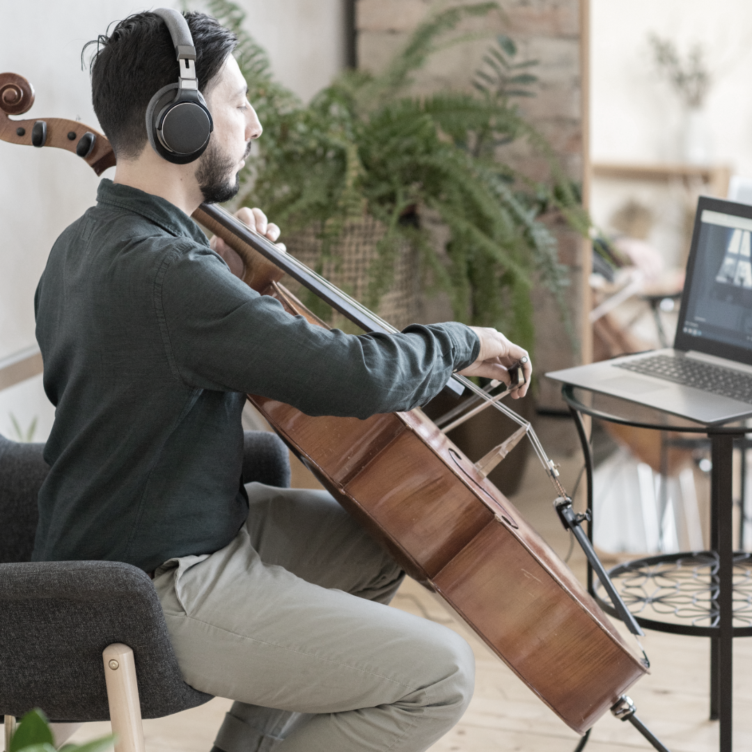 A man with headphones playing a cello while sitting on a chair in a room with plants and a laptop on a table.