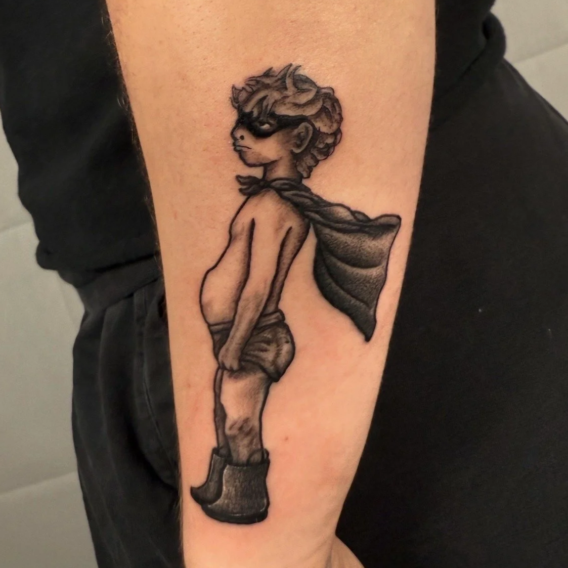 Tattoo of a child with curly hair, wearing sunglasses, a cape, shorts, and boots, standing with hands in pockets.