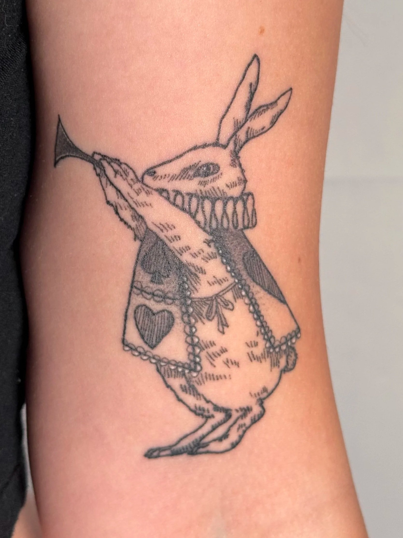 Tattoo of a rabbit with exaggerated teeth, holding a fish, wearing a dress with a heart on it, and a pearl necklace.