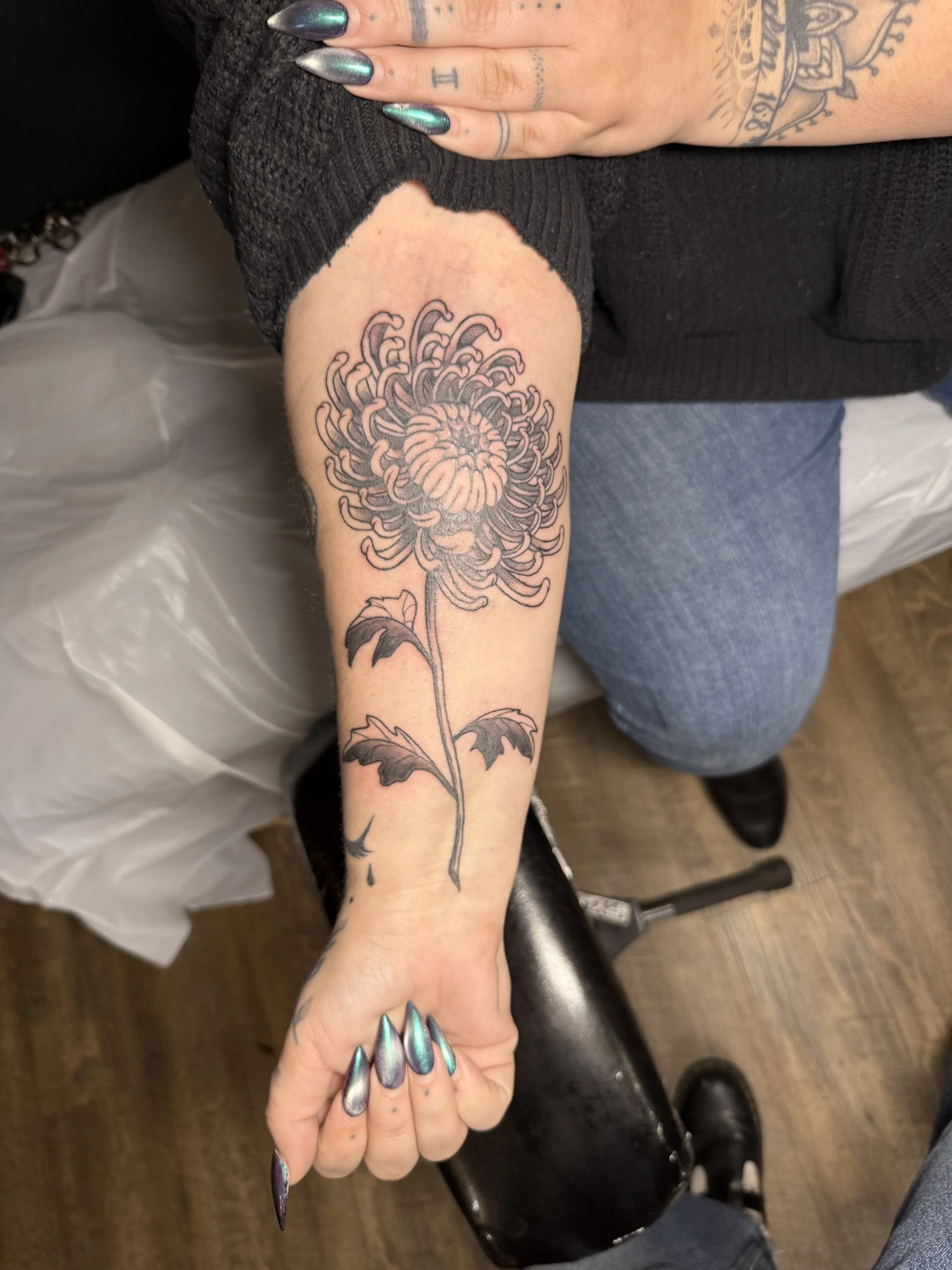 A person with metallic teal and purple nails shows their forearm tattoo of a large chrysanthemum flower with detailed petals and leaves.