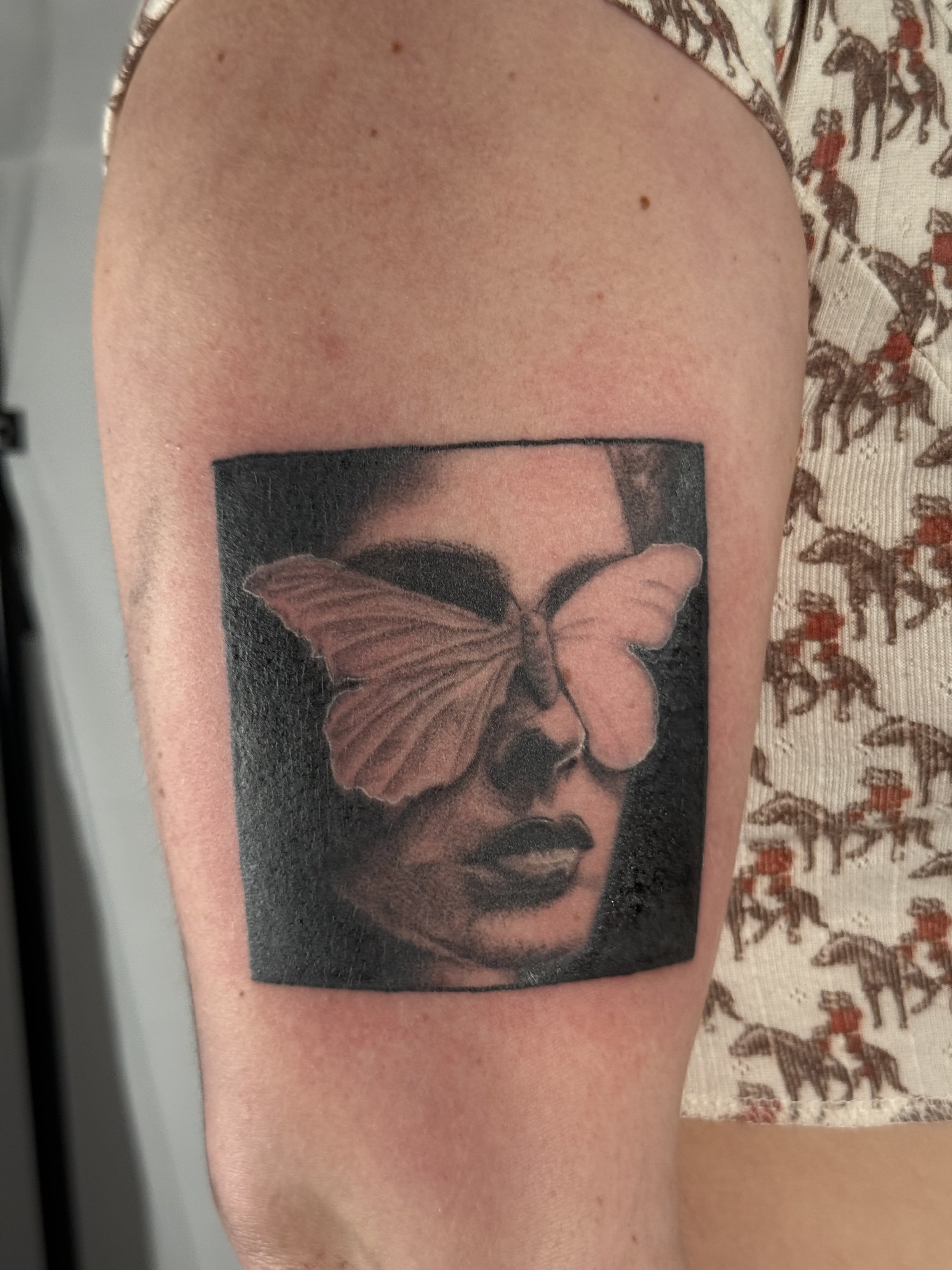 Tattoo of a woman's face with a large butterfly covering one eye, shaded in black and gray, on an arm.