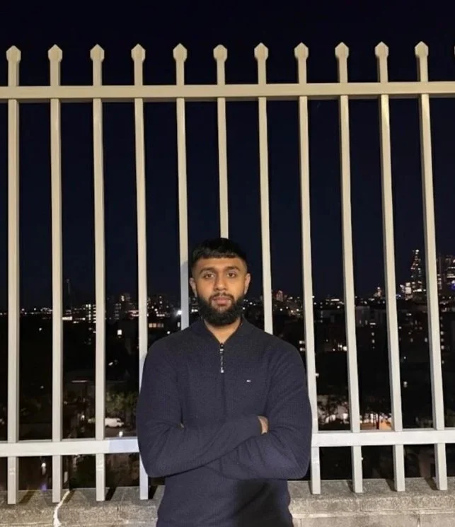 Young man with beard and short dark hair, standing with arms crossed in front of a metal fence at night, city skyline with illuminated buildings in the background.