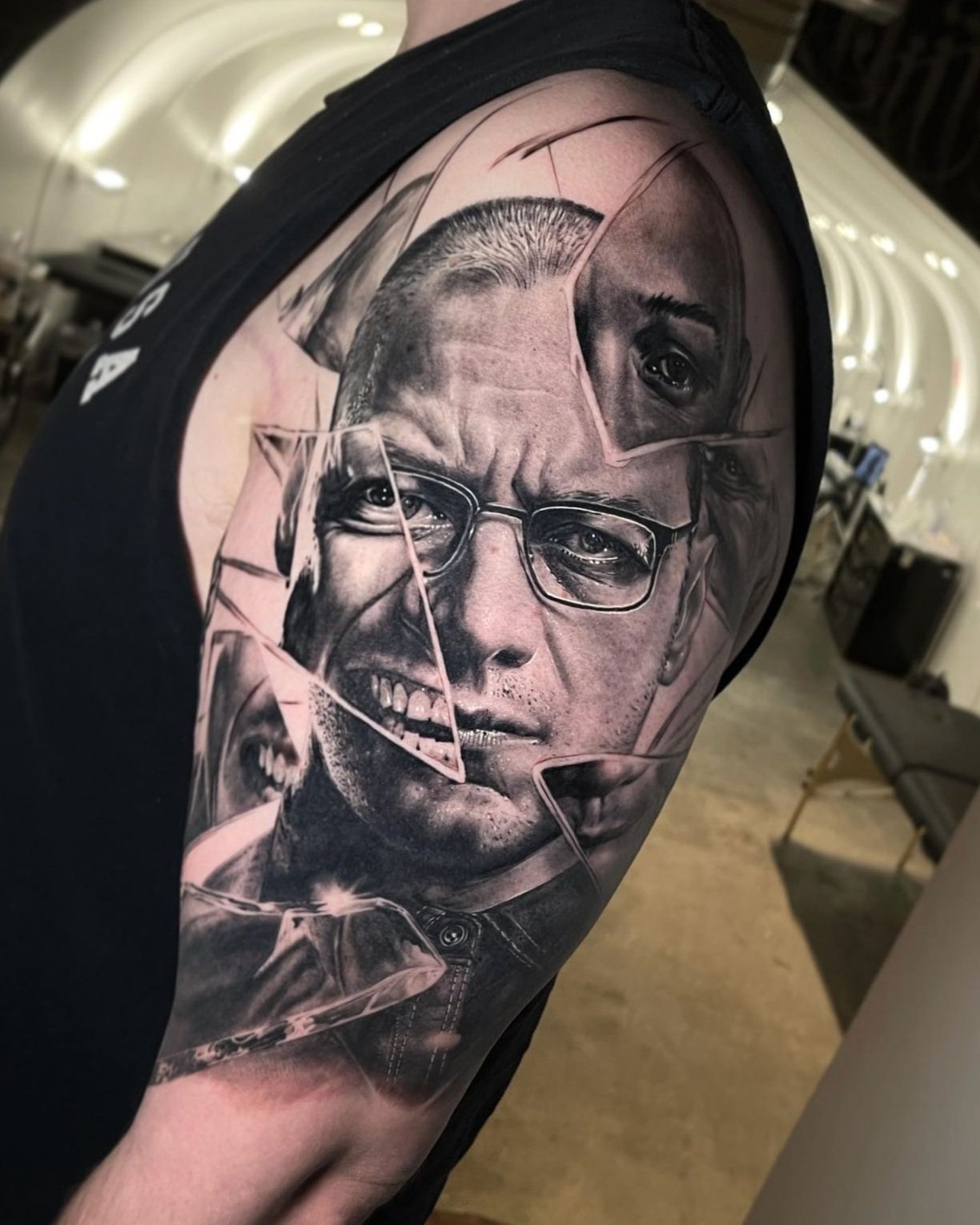 Close-up of a tattoo on a person's upper arm featuring a realistic black and gray portrait of a man with glasses, smiling, with a broken glass effect over the face in a tattoo studio setting.