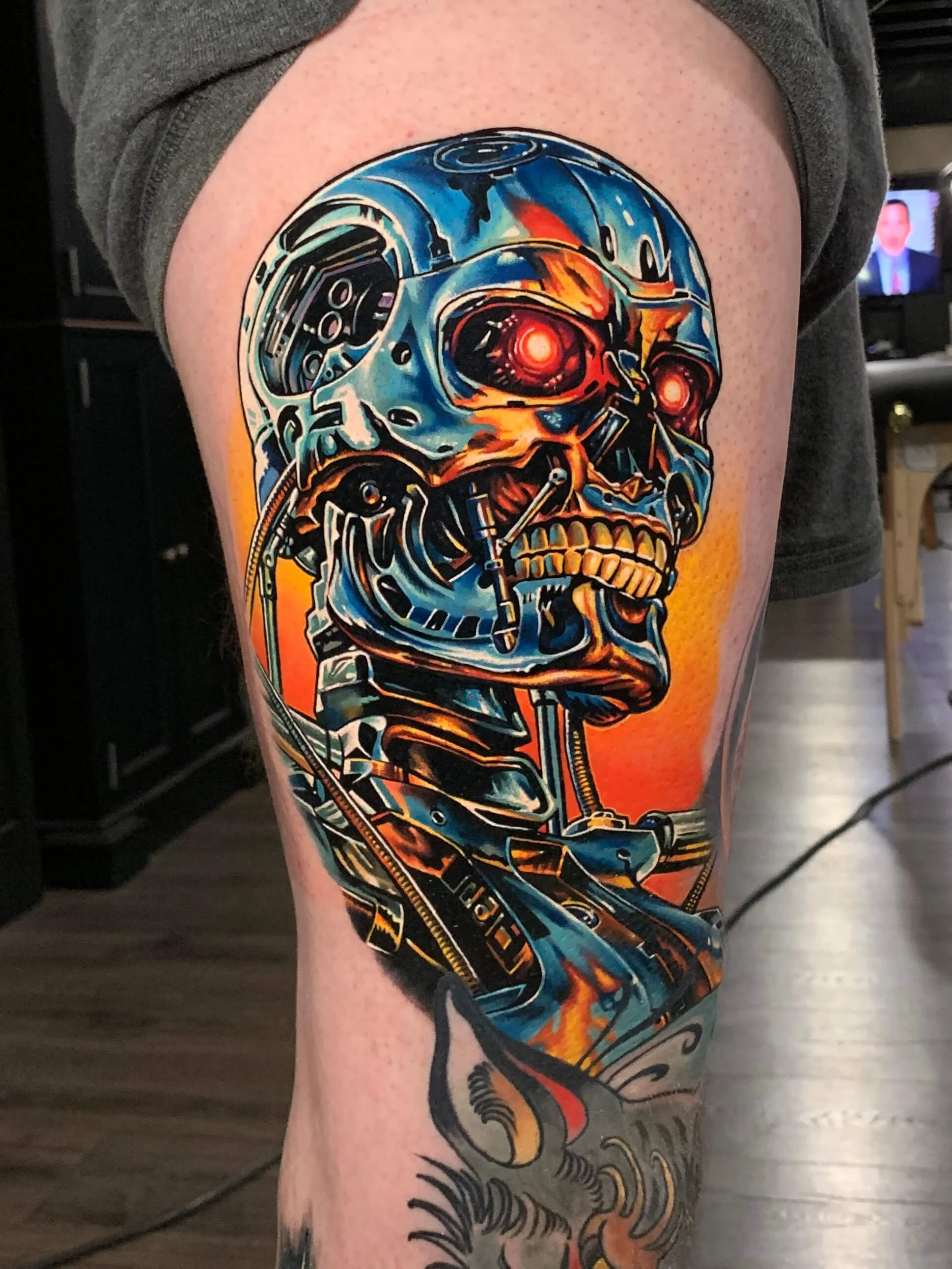 Colorful tattoo of a mechanical skull with glowing red eyes on a person's thigh.