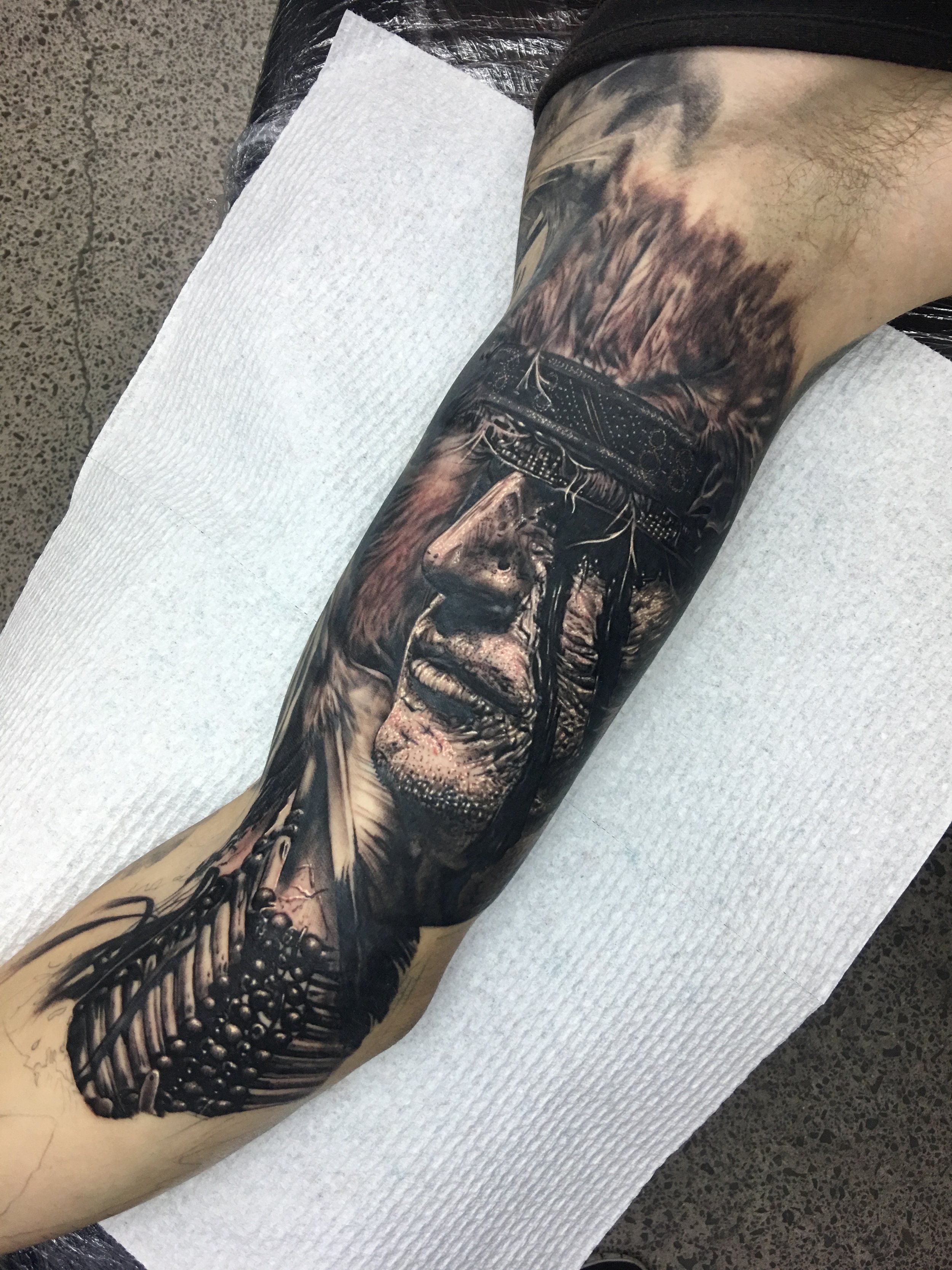 Close-up of a person's leg with detailed black and gray fantasy-style tattoo artwork featuring a face, feathers, and abstract patterns.