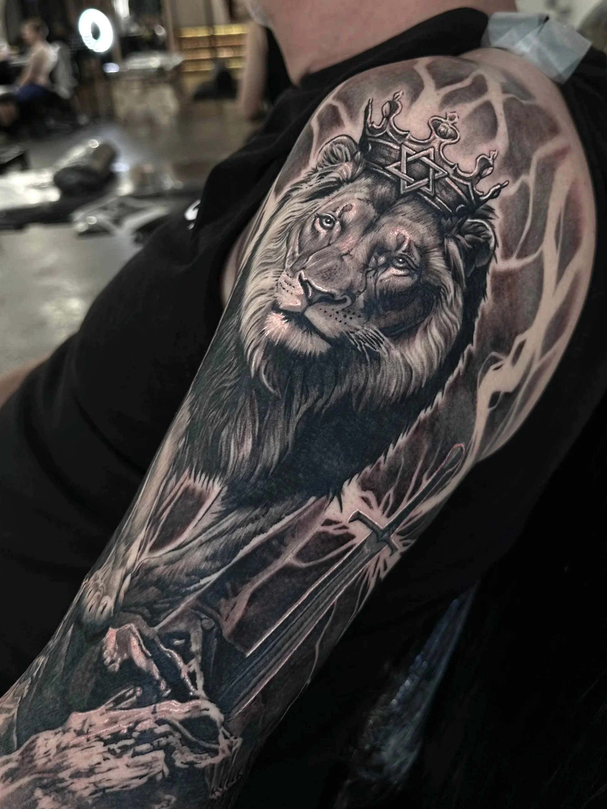 Close-up of a tattoo on a person's shoulder featuring a realistic lion wearing a crown, with intricate details and shading.