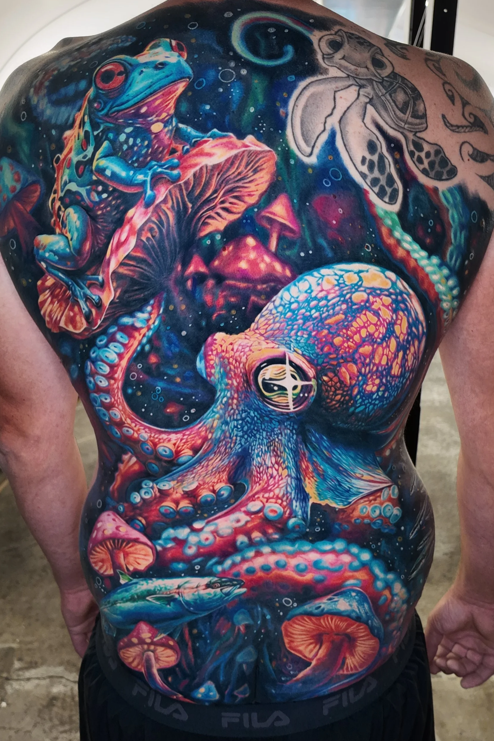 Colorful tattoo of a frog, mushrooms, and abstract cosmic elements covering the person's back and shoulder.
