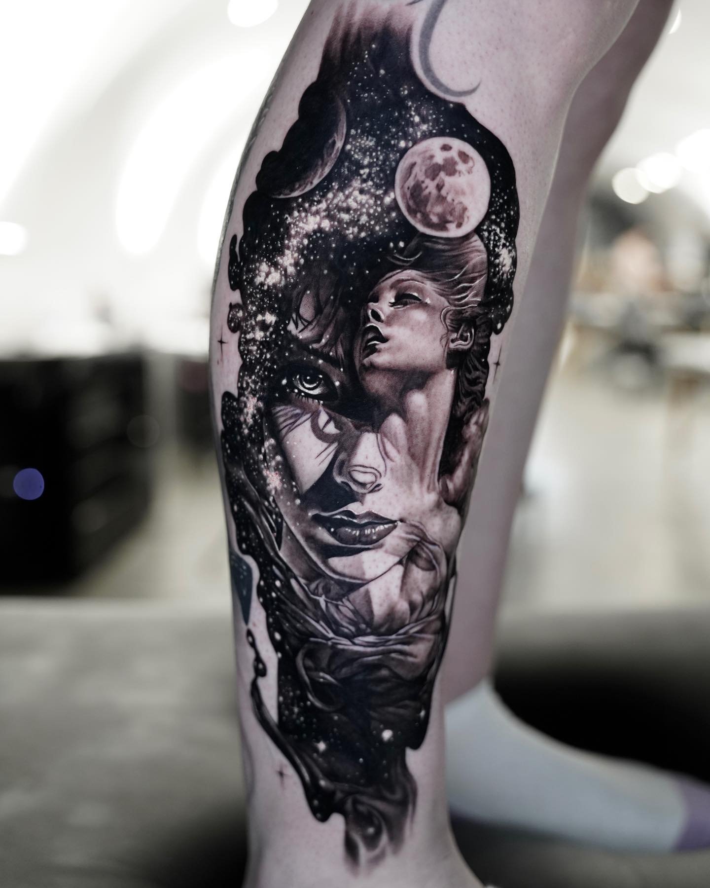 Tattoo of a woman's face with her eyes closed, surrounded by cosmic elements like moons and stars, in black and white ink.