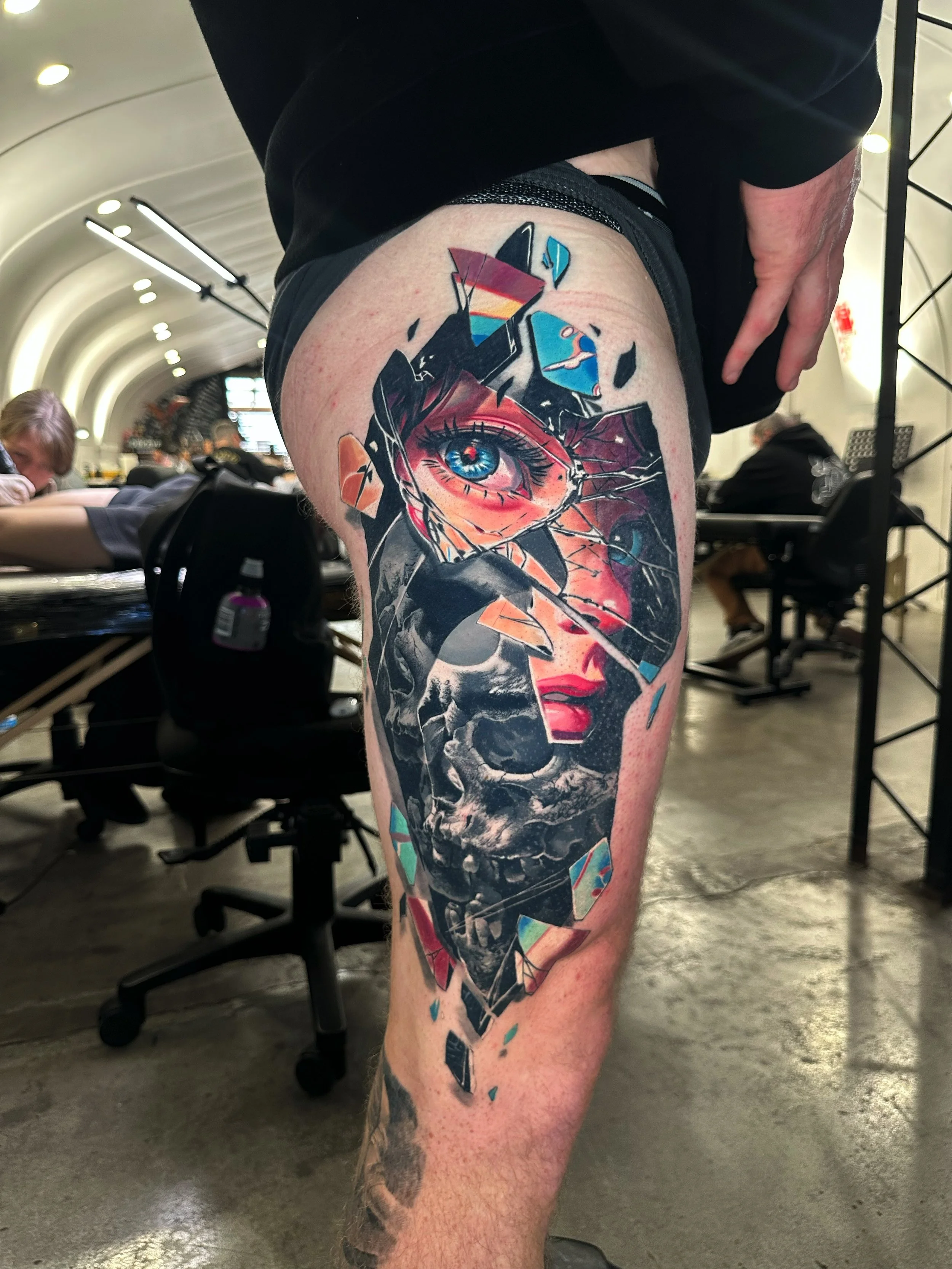 Tattoo of a shattered mirror reflection of a woman's eye and face on a person's upper thigh inside a tattoo parlor.