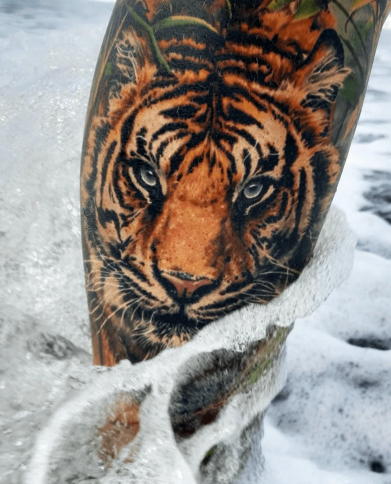 Close-up of a tattoo of a tiger's face on a person's arm, showing detailed orange, black, and white fur markings, with water and foam around the arm.