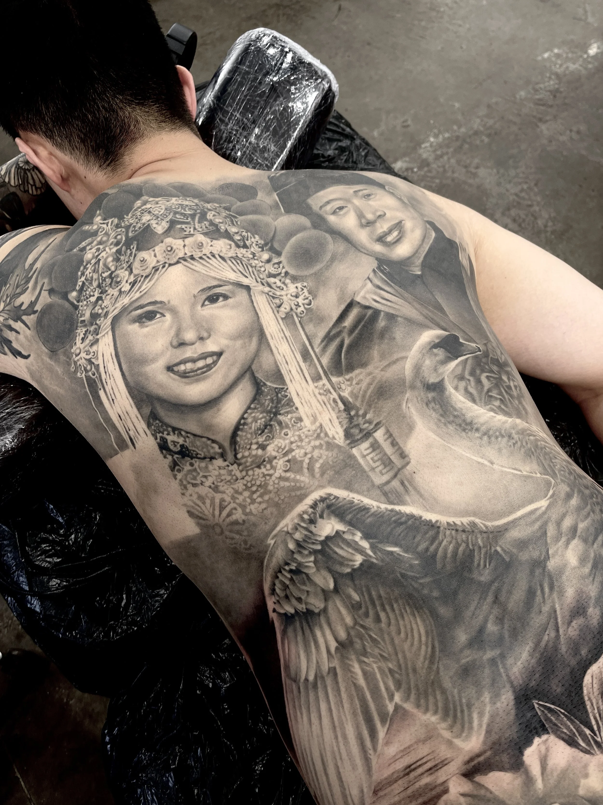 Black and white tattoo of a woman wearing traditional clothing and a headdress, and a man with short hair, on a person's back.