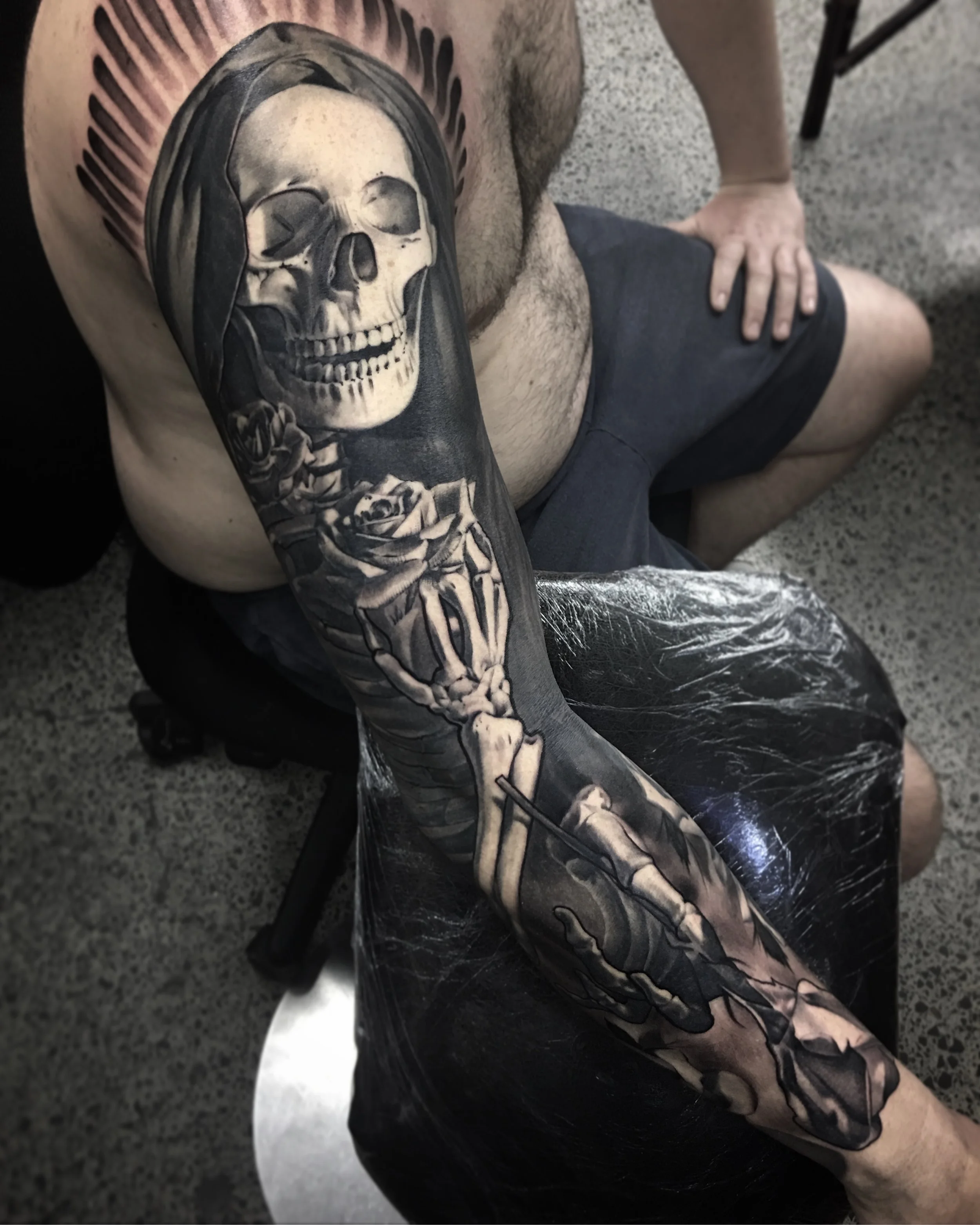 A person with a large, detailed black and gray tattoo sleeve depicting a skull, a playing card, and floral elements on their arm. The individual is sitting on a chair with their hand on their knee, wearing dark shorts and a shirtless upper body, on a