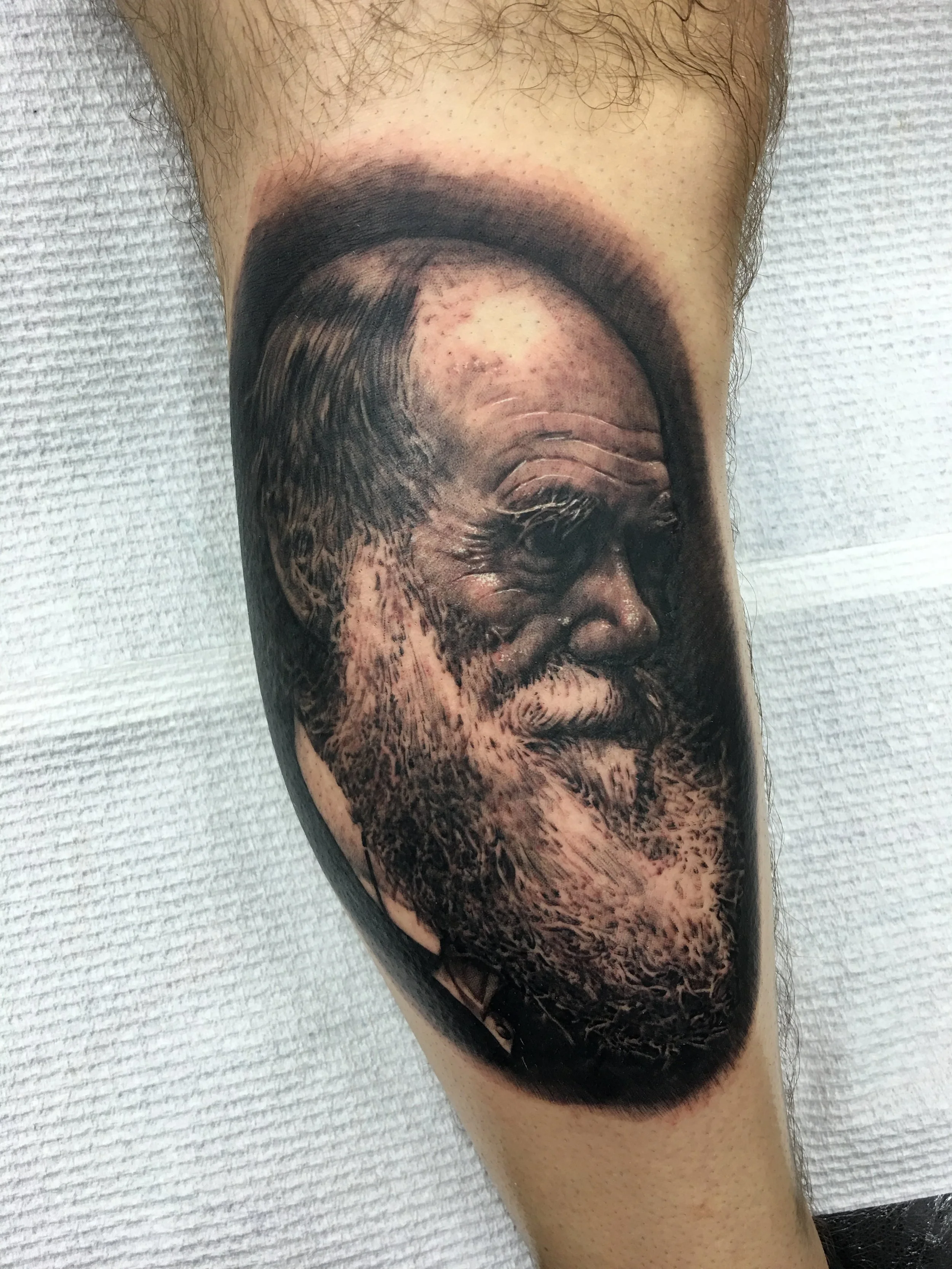 Tattoo of a man's face with closed eyes, full beard, and long hair, shaded in black and gray realism style, covering most of the person's calf.