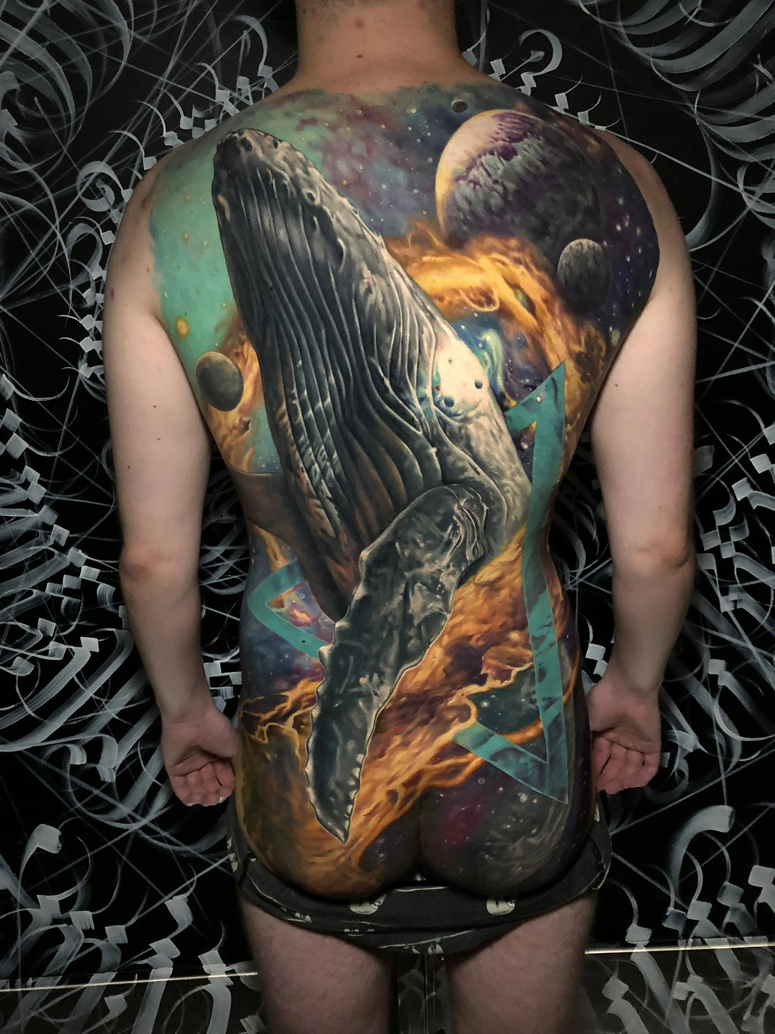 A person's back and upper buttocks painted with a space-themed mural, featuring planets, stars, and cosmic colors.