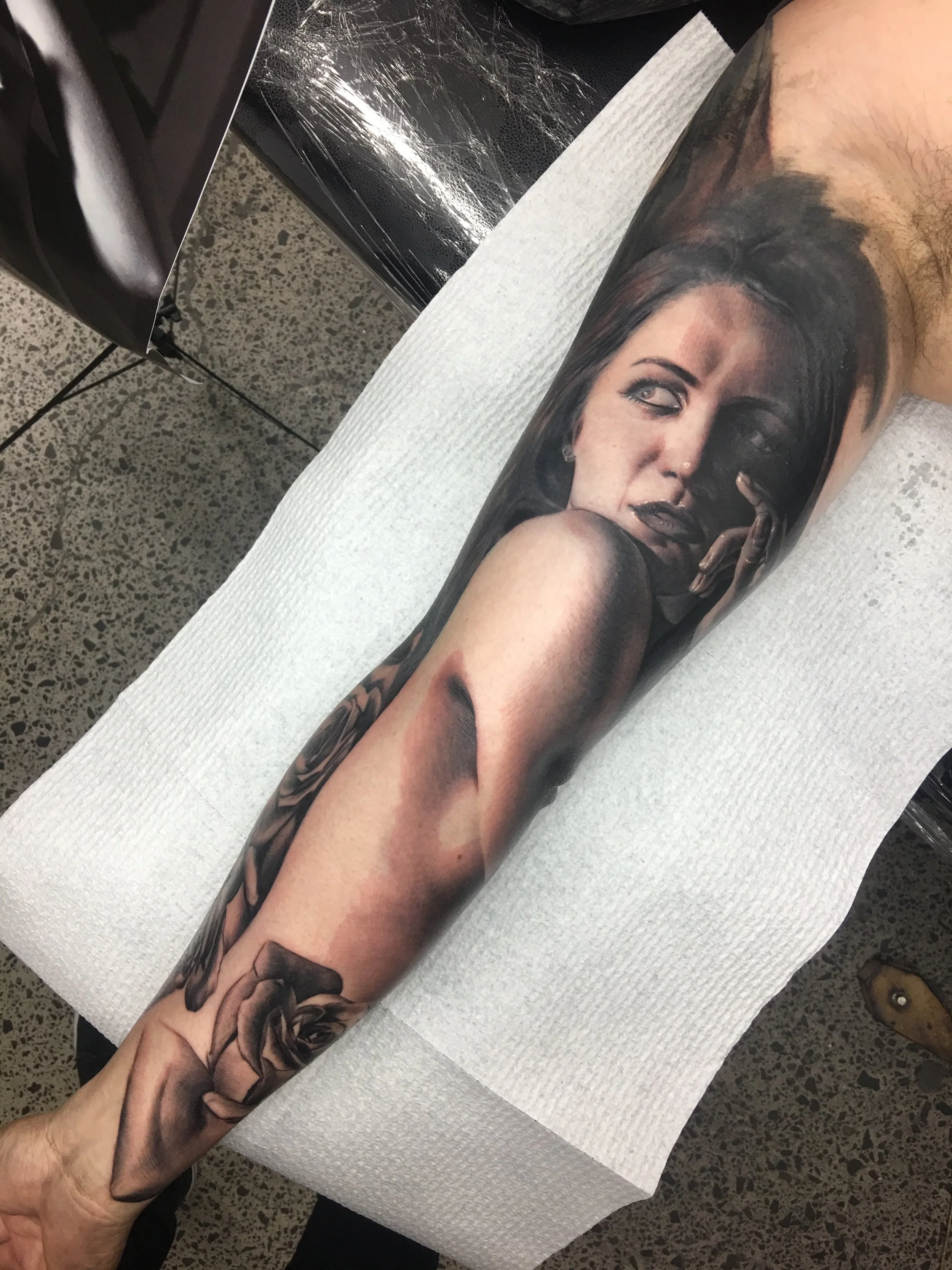 A person with a detailed black and gray tattoo sleeve on their arm, lying on a white paper-covered surface at a tattoo studio, with part of their torso and face visible in the tattoo.