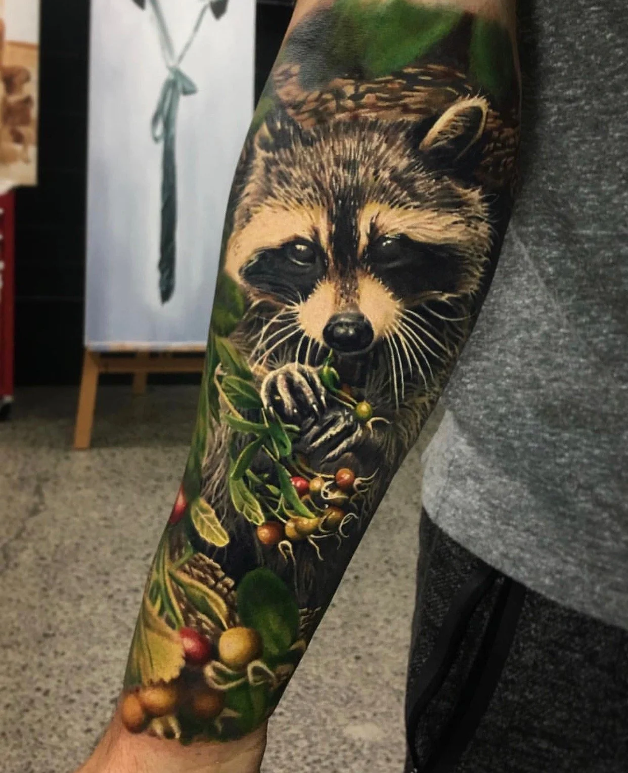 Tattoo of a raccoon holding a plant branch with berries and leaves on a person's arm.