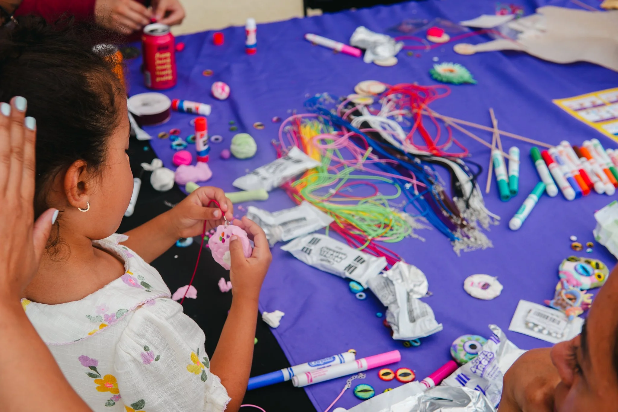 A young girl is engaged in a crafting activity at a table covered with colorful craft supplies, including yarn, glue sticks, googly eyes, and foam shapes.