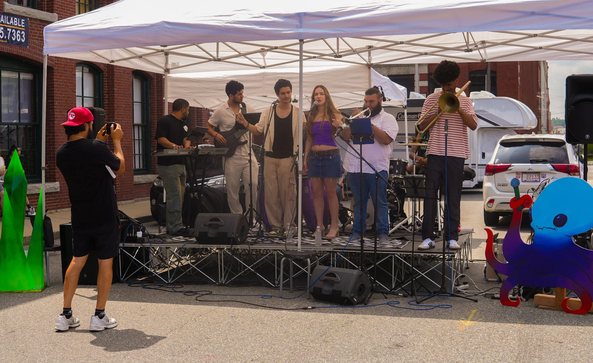A group of musicians performing on a small outdoor stage under a white canopy tent, with a man in a pink cap taking photos, and colorful playful octopus and sword decorations nearby.