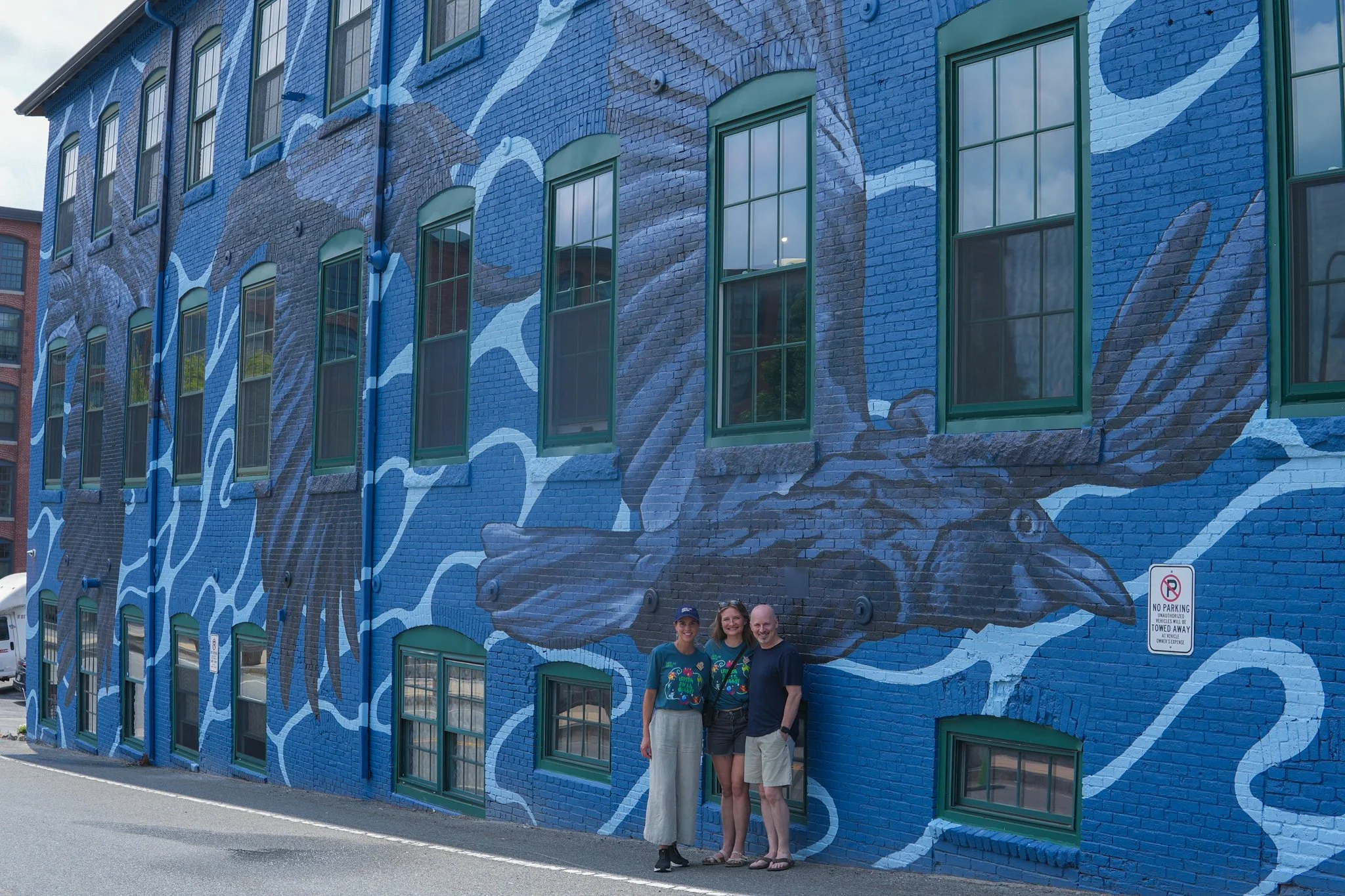 Three people standing in front of a large blue mural painted on a brick building, depicting a giant squid with tentacles extending across the wall.