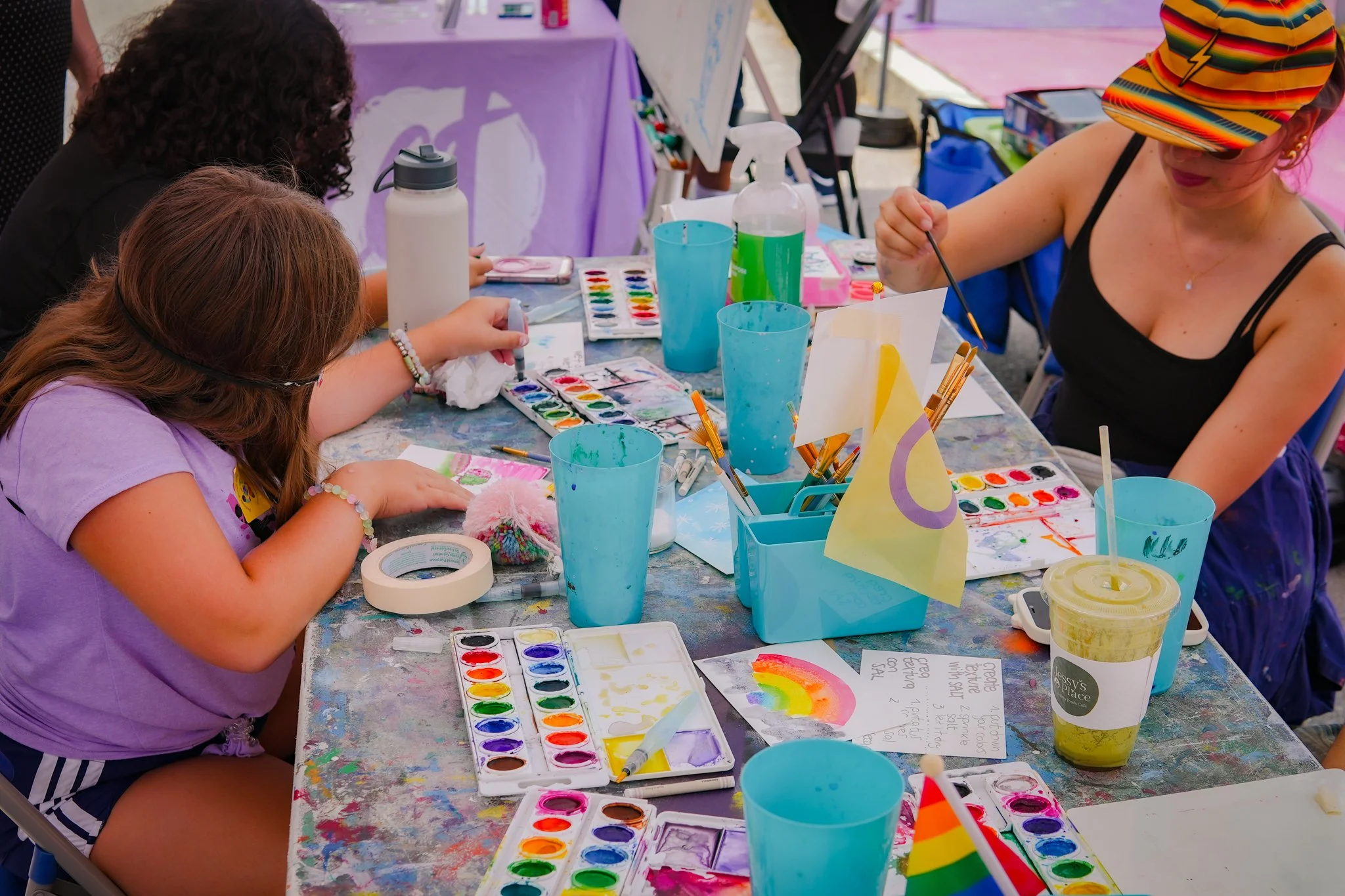 Children and an adult painting watercolors at a colorful table during an outdoor art activity, with supplies including watercolors, paintbrushes, cups, and papers.
