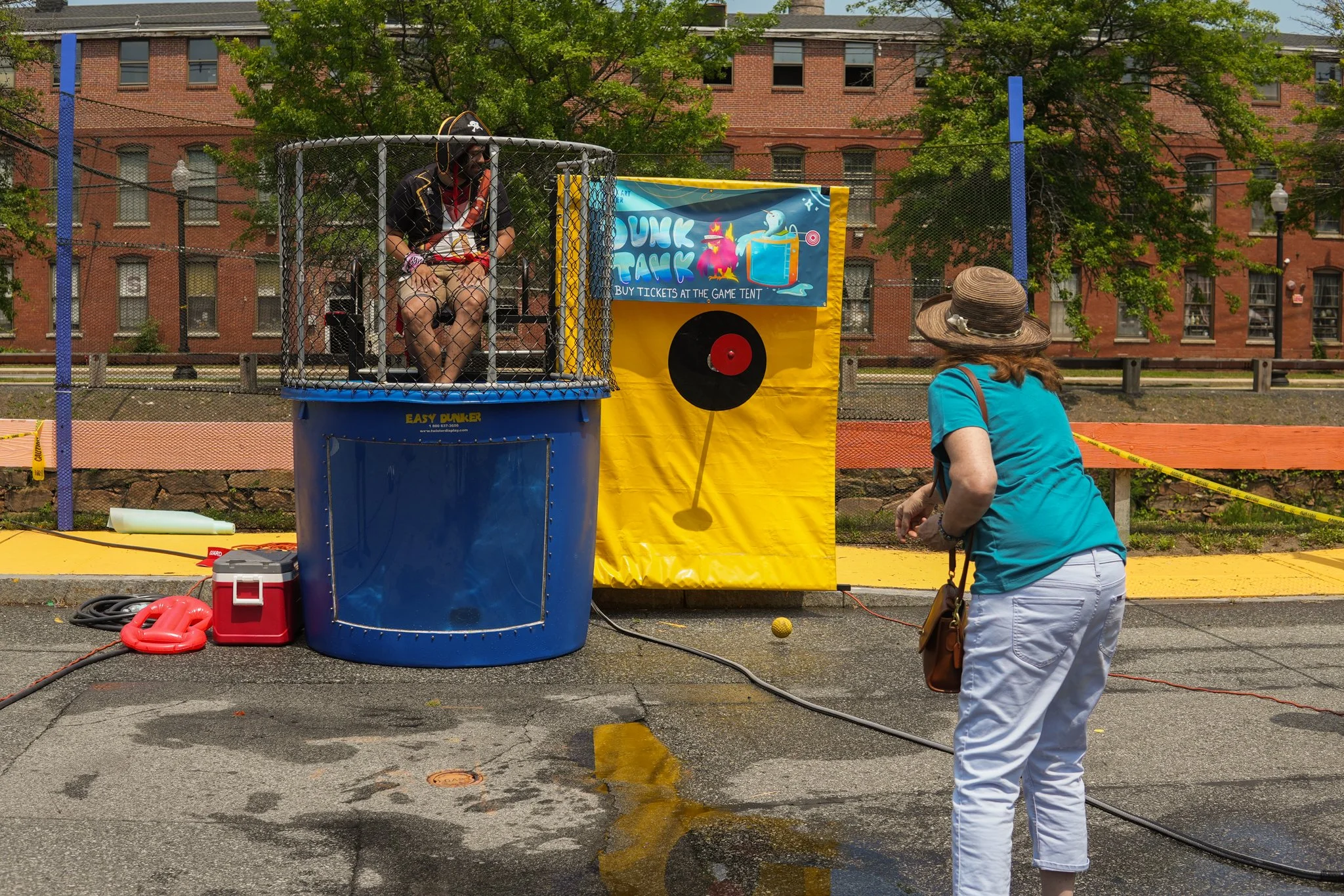 A woman in a turquoise shirt and white pants is aiming a water gun at a person sitting in a dunk tank. The person in the tank is wearing a pirate hat and sitting on a seat with a mechanical arm, ready to be dunked. The dunk tank is blue with a chain-