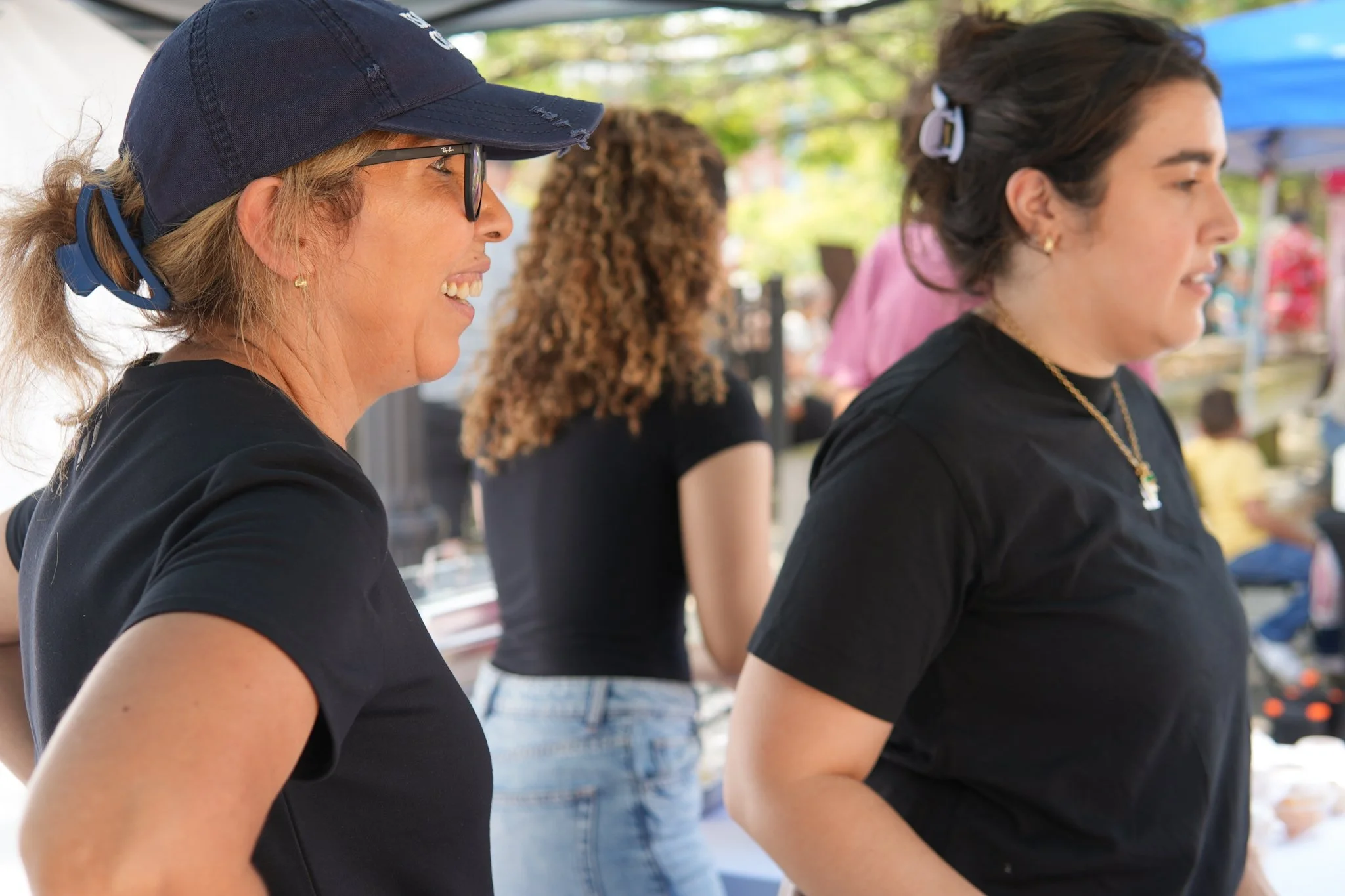 Two women wearing black shirts and glasses, standing and smiling outdoors at a market or fair.