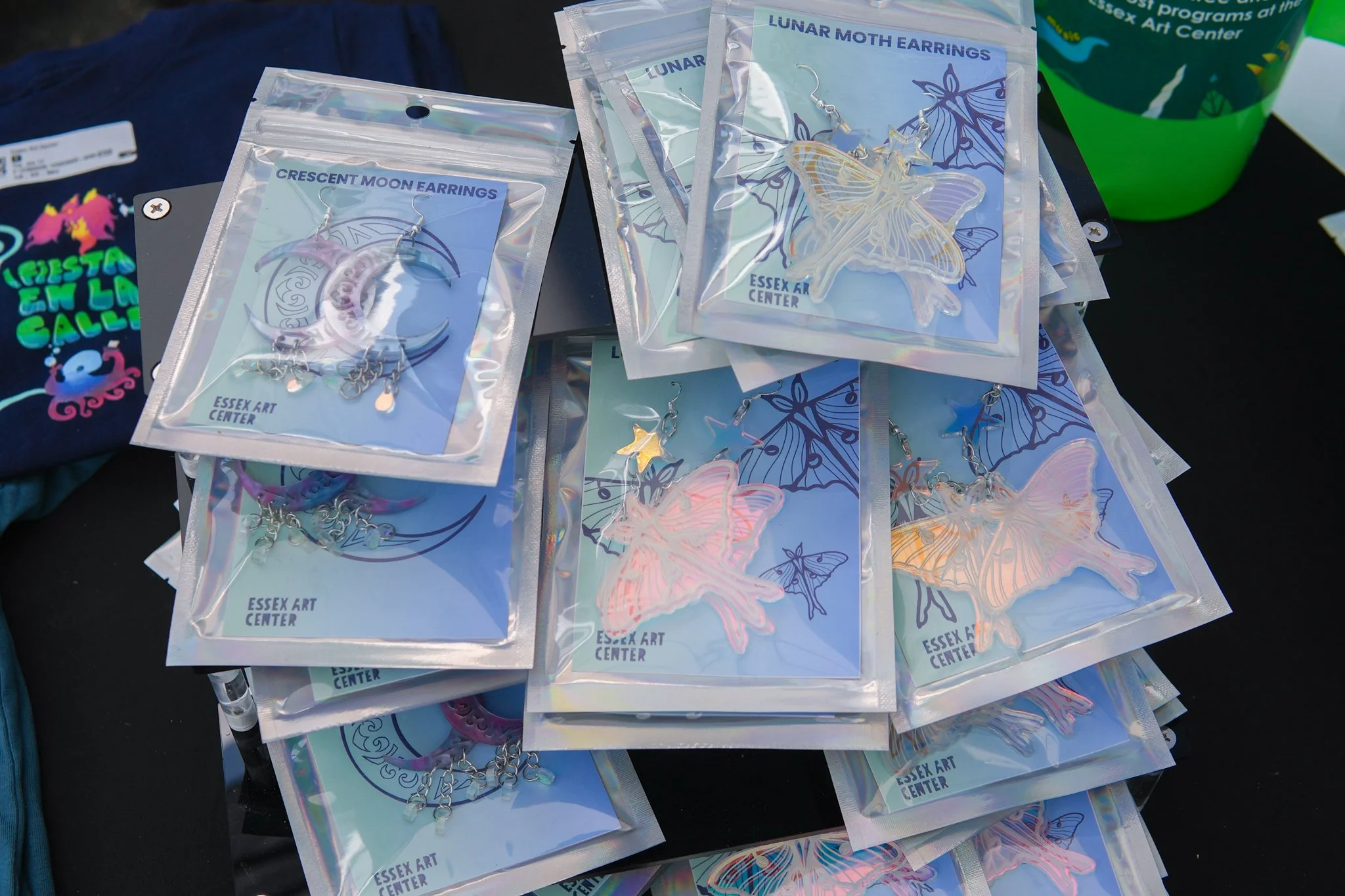 Multiple packages of earrings featuring lunar moth and crescent moon designs, labeled 'Essex Art Center', displayed on a black surface at an outdoor market.