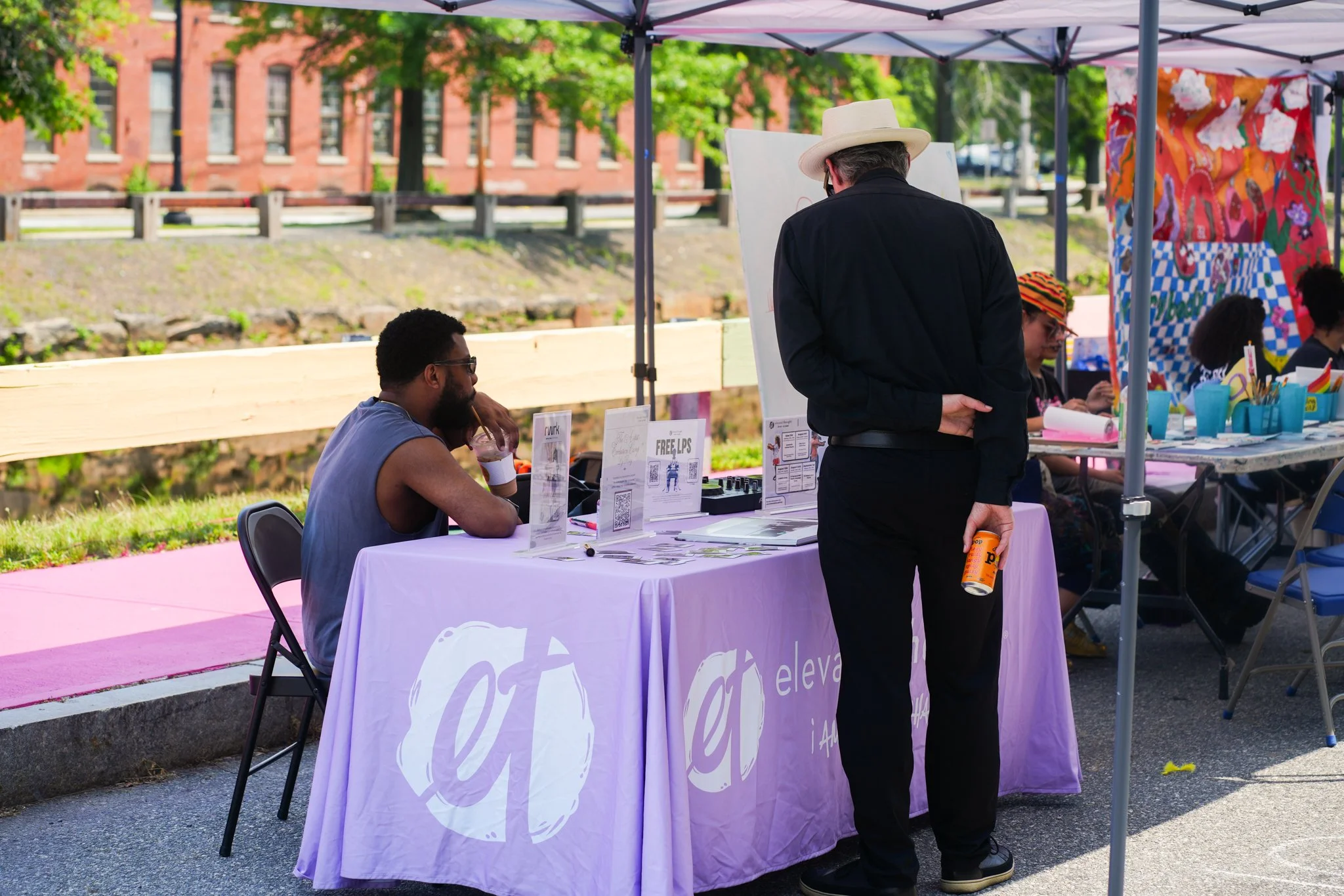 People sitting at a table with purple tablecloths at an outdoor event, engaging in conversation. One person is standing, holding a container, wearing a light-colored hat and black outfit. The booth has informational displays and promotional materials