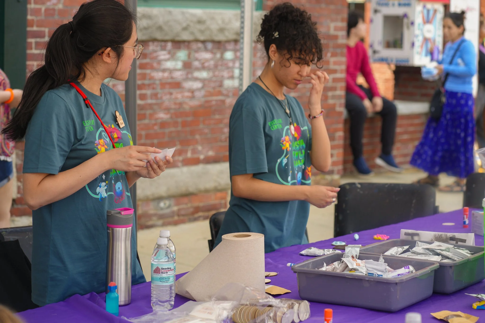 Two young women at a table outdoors, organizing items for an event, with one holding a small paper and the other touching her nose, dressed in dark teal t-shirts. In the background, other people are sitting on a brick wall and standing near a booth.