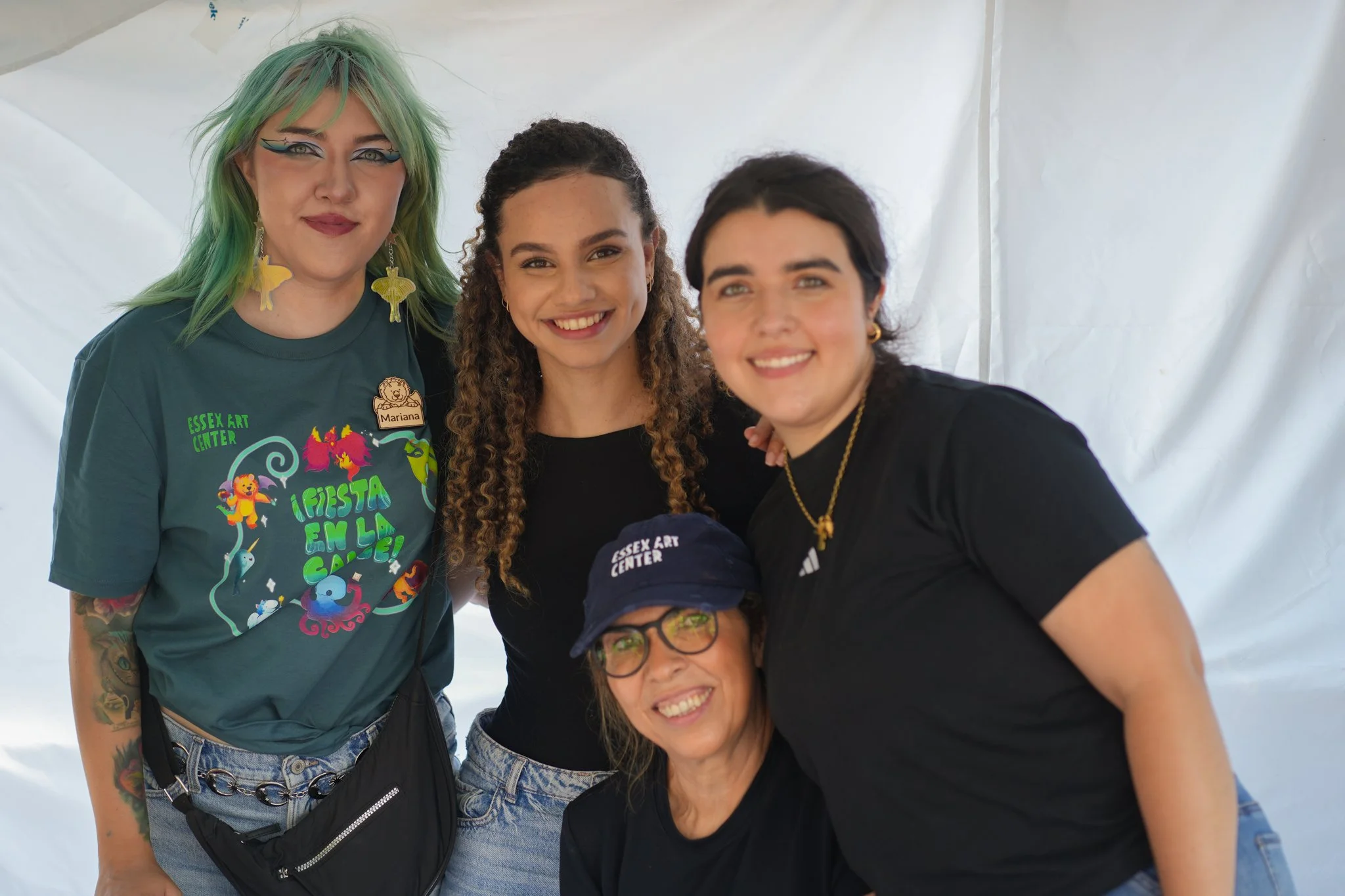 Four women smiling together inside a white tent at an event, one wearing a teal T-shirt with colorful graphics and a name tag, another in black, and one in a black T-shirt with a NASA logo. The woman in front is wearing glasses, a navy baseball cap,