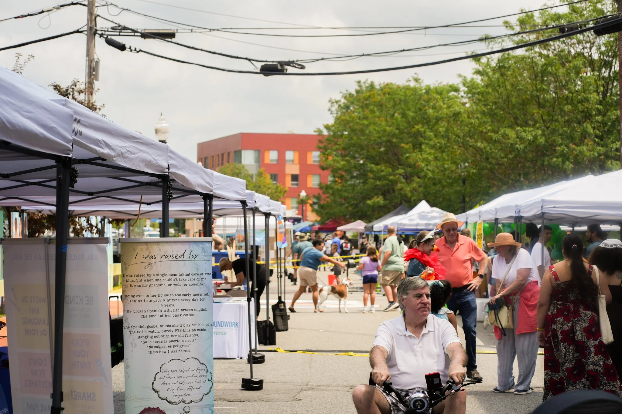 A street market with white tents and people walking, some with dogs, in a city setting with trees and a red brick building in the background.