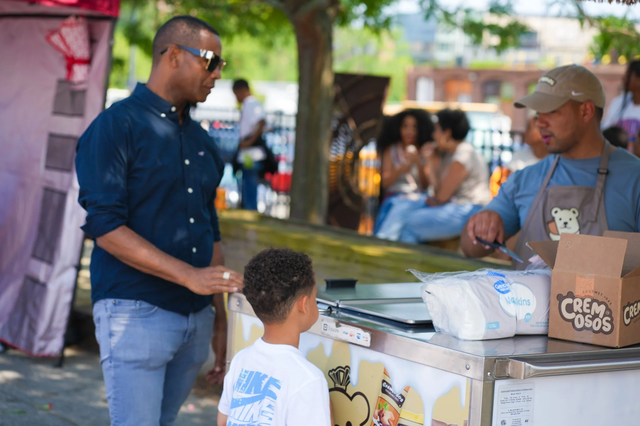A man in a dark blue shirt and sunglasses orders ice cream at a street stand, where a young boy in a white Nike shirt is also looking at the menu. The ice cream stand has a box of Cone-O-Sos and a bag of Wilkins marshmallows on the counter, with two