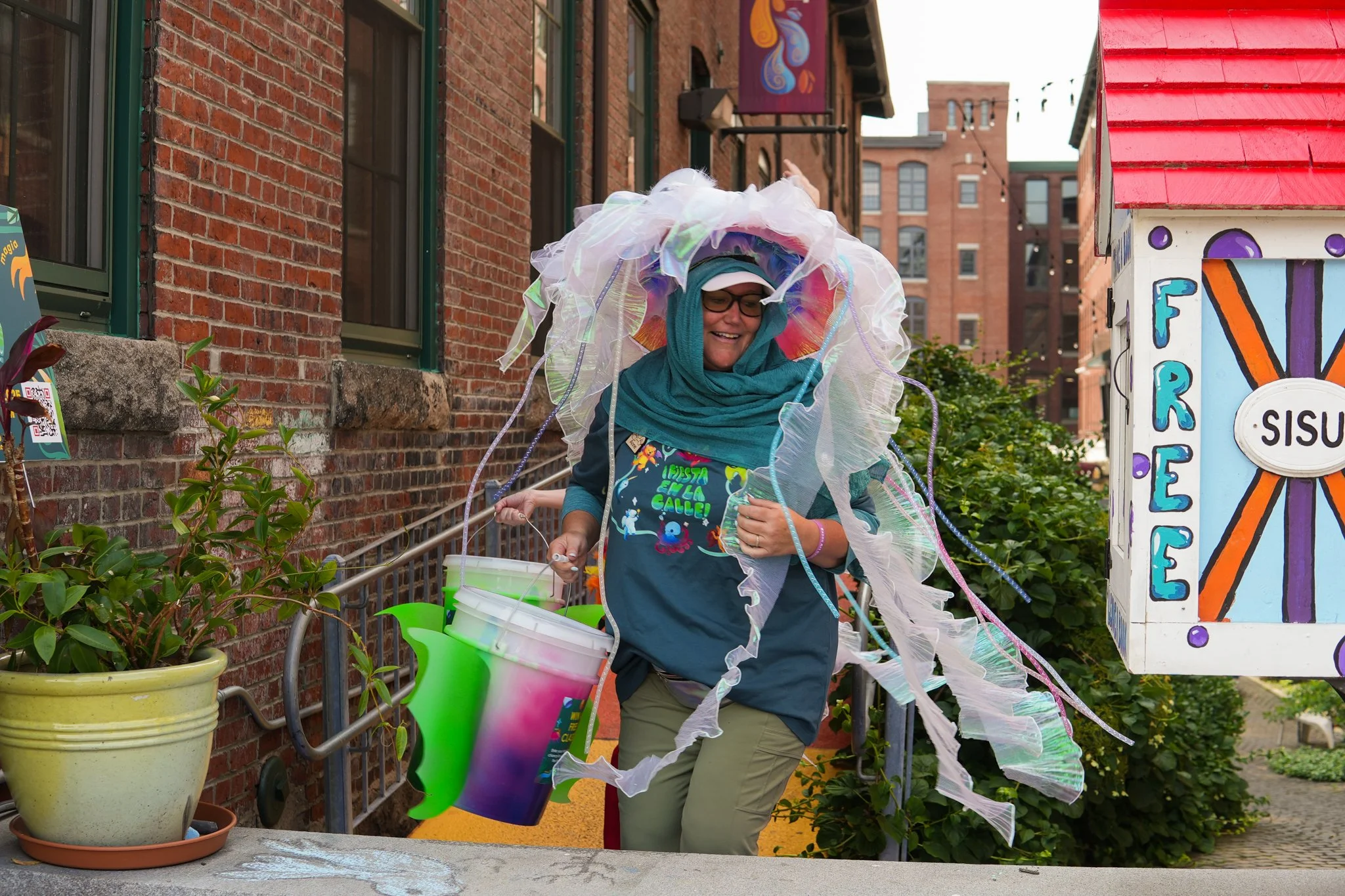Woman dressed as a mermaid, holding a colorful bucket, and walking outdoors on a city street, with a brick building and colorful sign nearby.