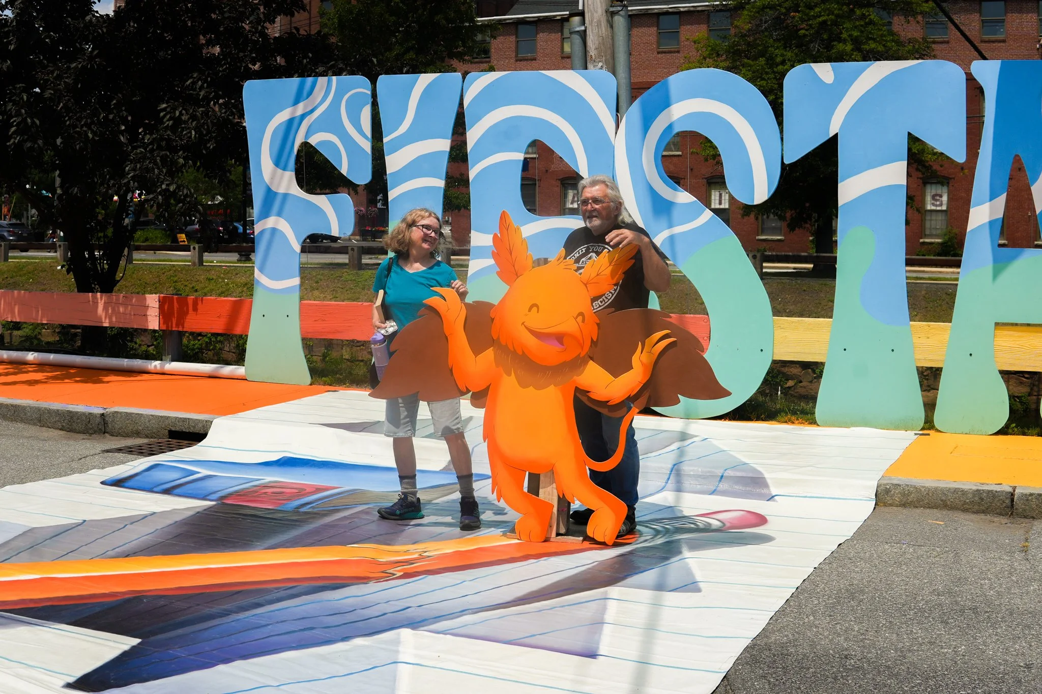 Two people, one young girl and one older man, stand behind a colorful cartoon lion mascot in front of large decorative letters spelling 'FLOS'. The scene appears to be outdoors on a sunny day with trees and buildings in the background.