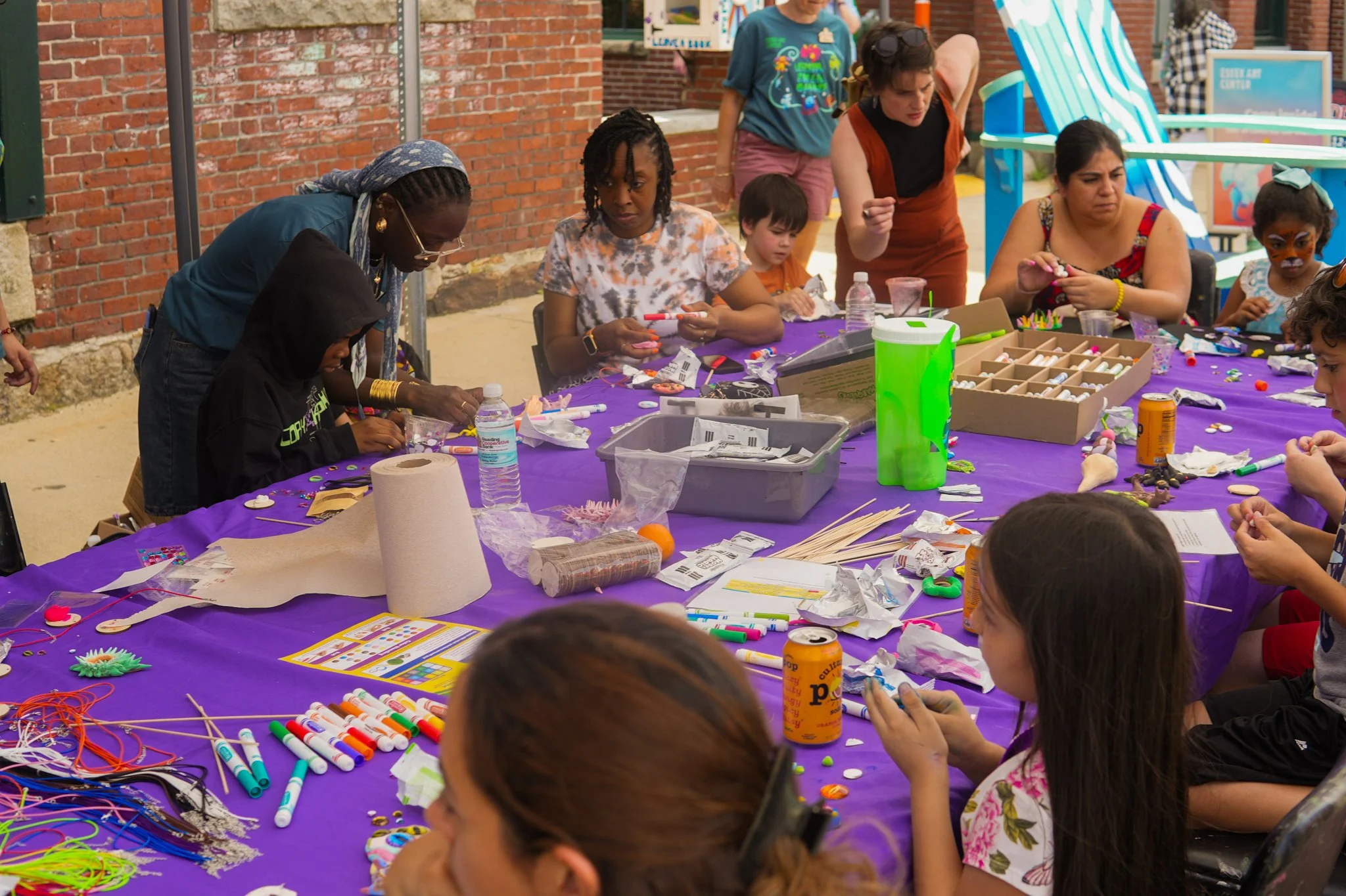 A group of children and adults sitting at a purple table outdoors, engaged in arts and crafts activities with various supplies like markers, glue, paper, and decorations.