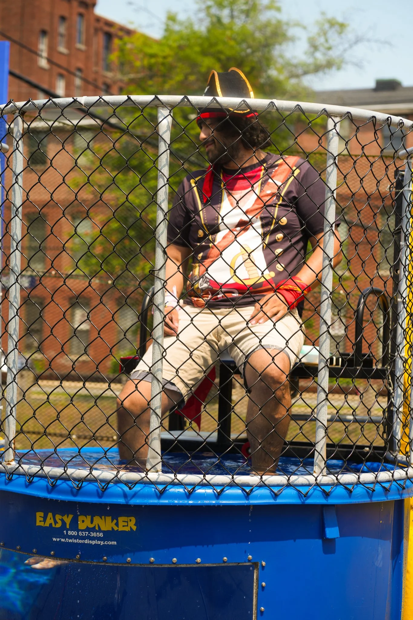 A man dressed as a pirate sitting on a chair inside a water dunk tank with a blue base labeled "EASY DUNKER" and a chain-link enclosure, outdoors with trees and brick buildings in the background.