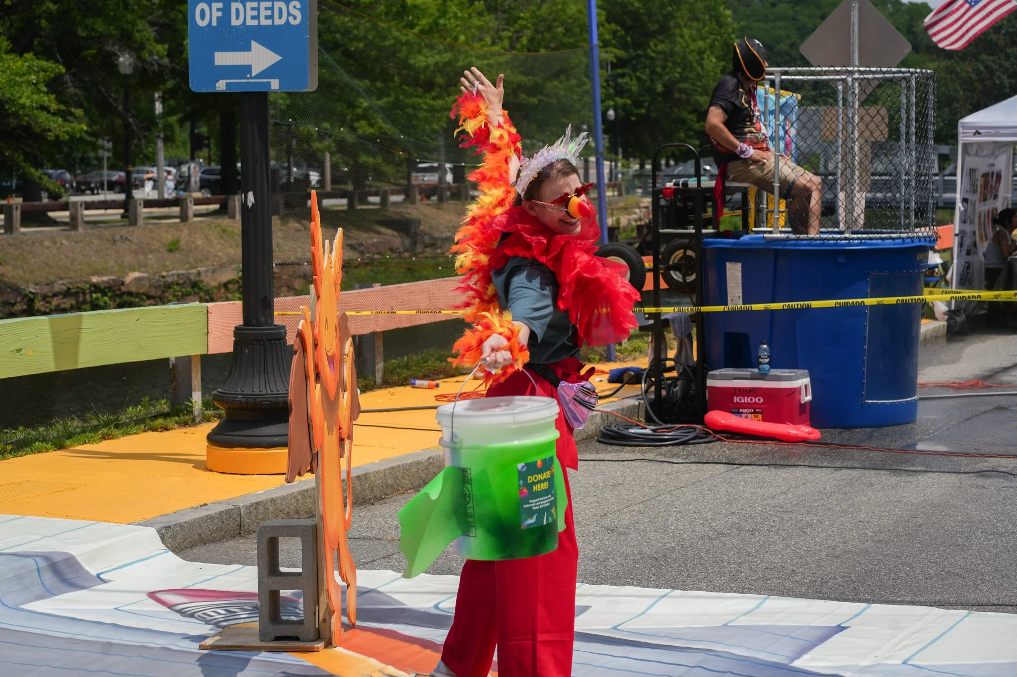 A person dressed as a clown with a red feather boa, clown nose, and angel halo stands on a street during a parade or festival, smiling and waving. They are holding a bucket labeled "Donate Here" and wearing red pants. Behind them, there is a hot air