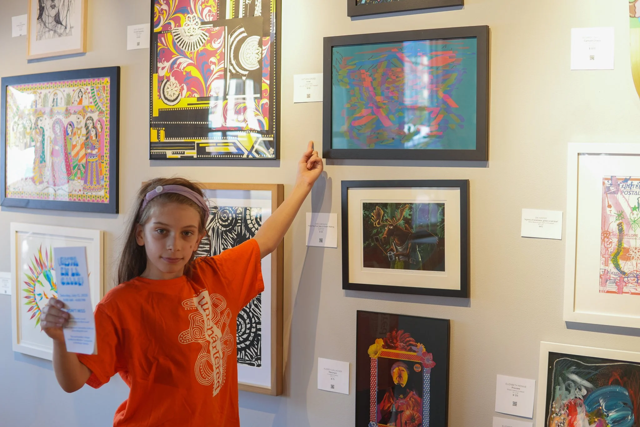 A young girl in an orange shirt points at colorful abstract artwork in an art gallery, holding a pamphlet in her other hand.