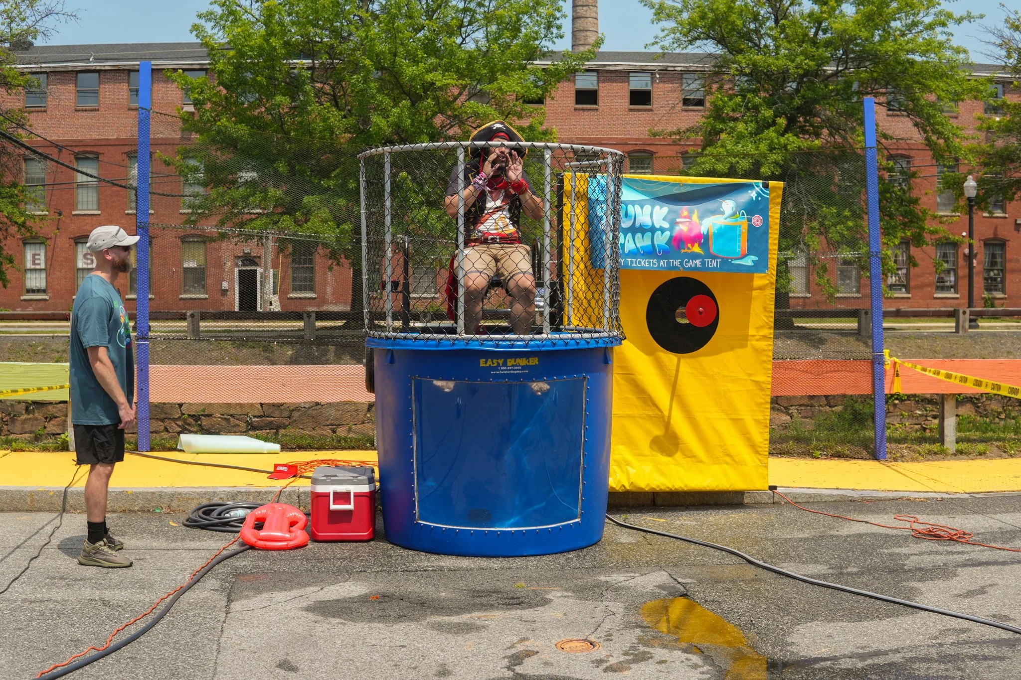 A person sitting inside a dunk tank, getting sprayed with water, while another person watches nearby. The dunk tank is set up outdoors with a yellow backdrop and a sign that says 'Dunk Tank'.