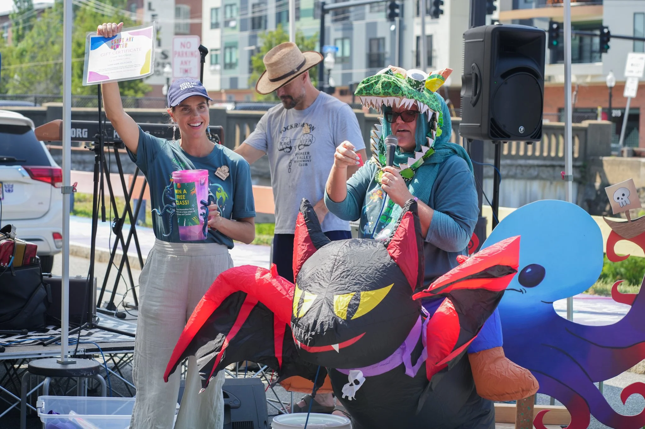 A woman holding a gift certificate smiling in front of a man and a woman dressed in a dragon costume with red and black wings, green spikes, and a skull decoration around the neck, at an outdoor event.