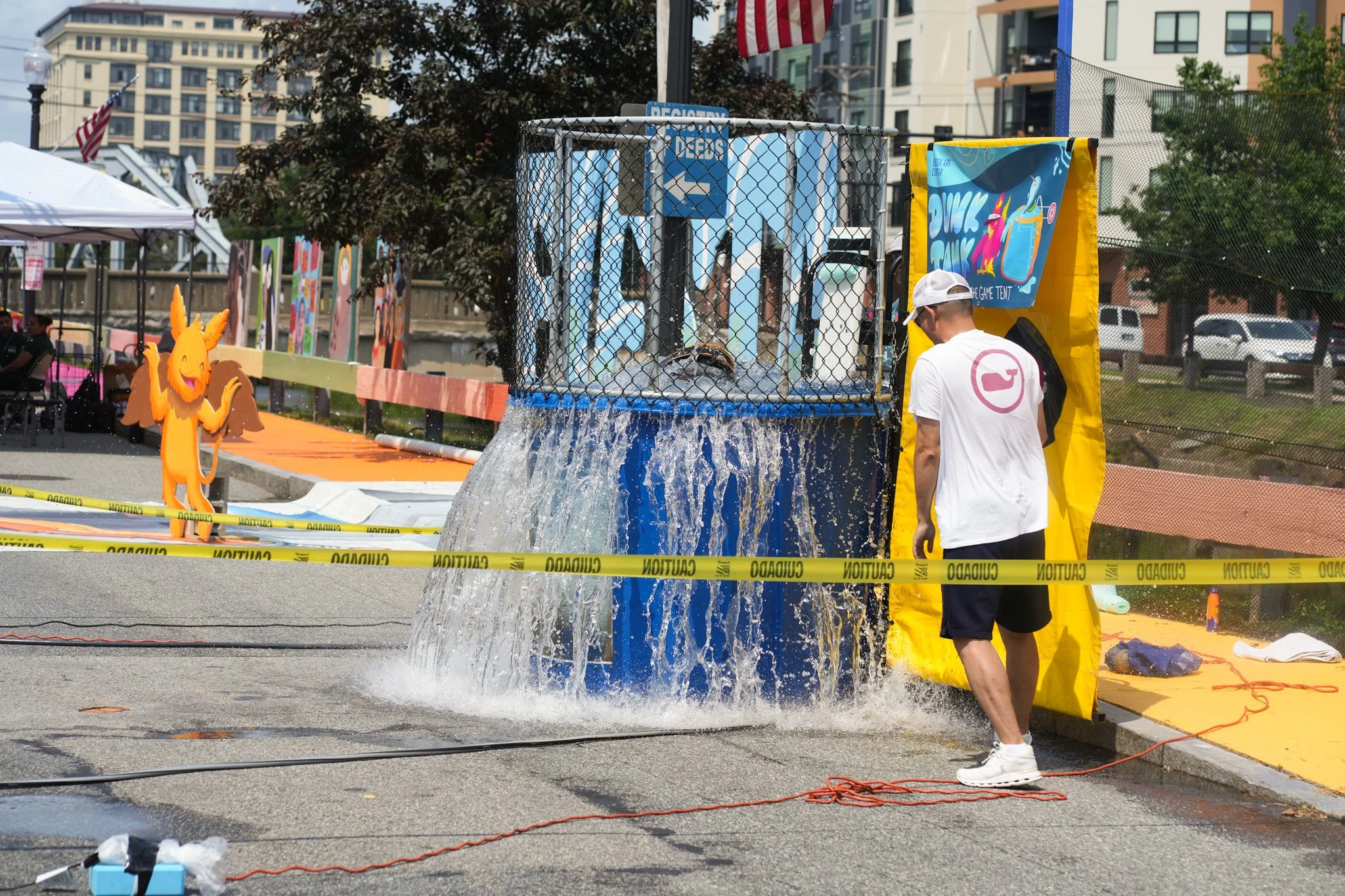 A person in a white shirt and shorts is standing under a water fountain at an outdoor event, with caution tape surrounding the area and colorful decorations and advertisements in the background.