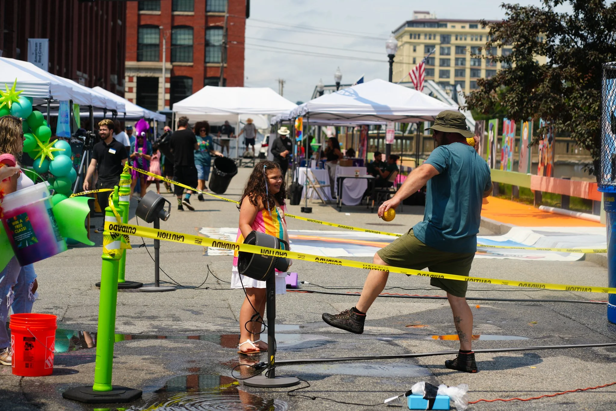 A young girl and an adult man playing a water game at an outdoor festival, surrounded by tents, balloons, and other attendees.