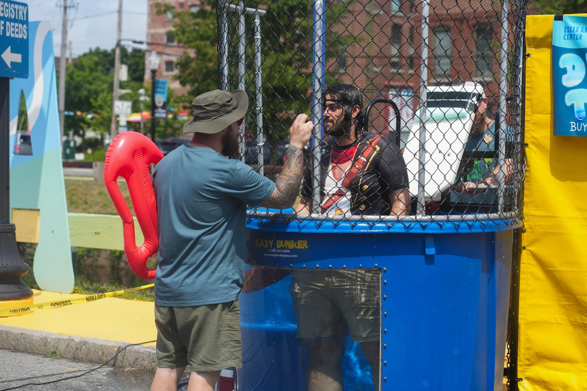 A man with a beard, wearing a blue t-shirt and a wide-brimmed hat, stands outside a dunk tank, inflating a red balloon to dump water on a man inside the tank. The man inside the tank has glasses, a beard, a black shirt with a colorful design, and is