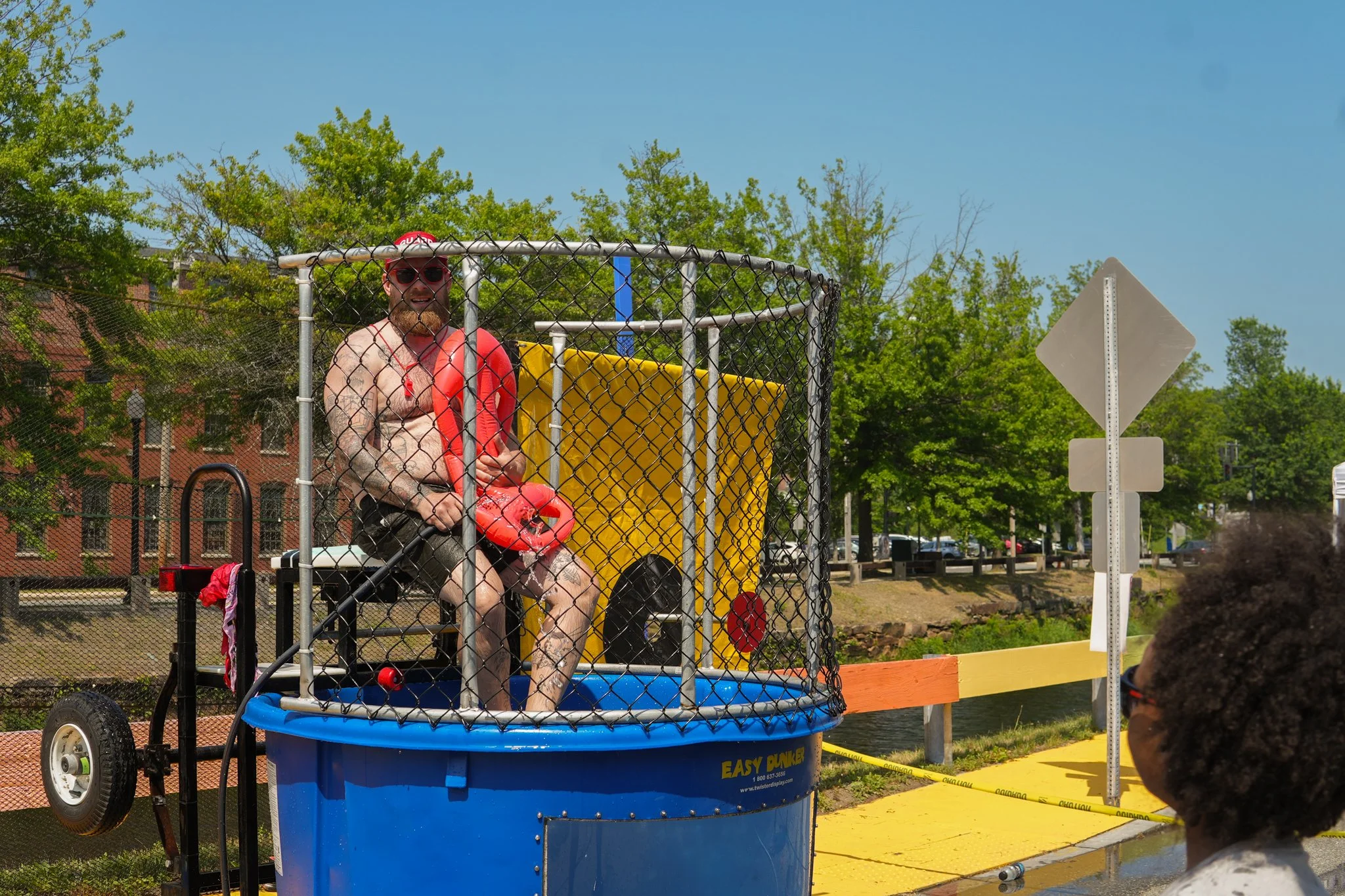 Man sitting inside a dunk tank with a red life jacket, wearing sunglasses, and smiling. Person with curly hair and glasses is watching, in an outdoor park setting with trees and buildings in the background.