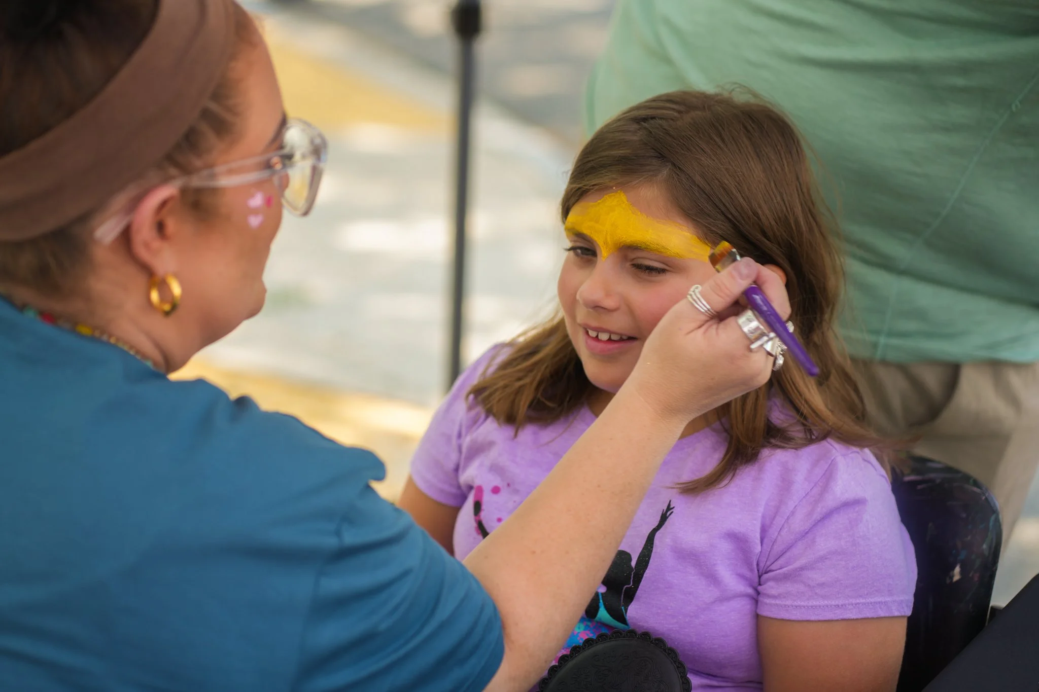 A woman is painting a yellow design on a young girl's forehead at an outdoor event.